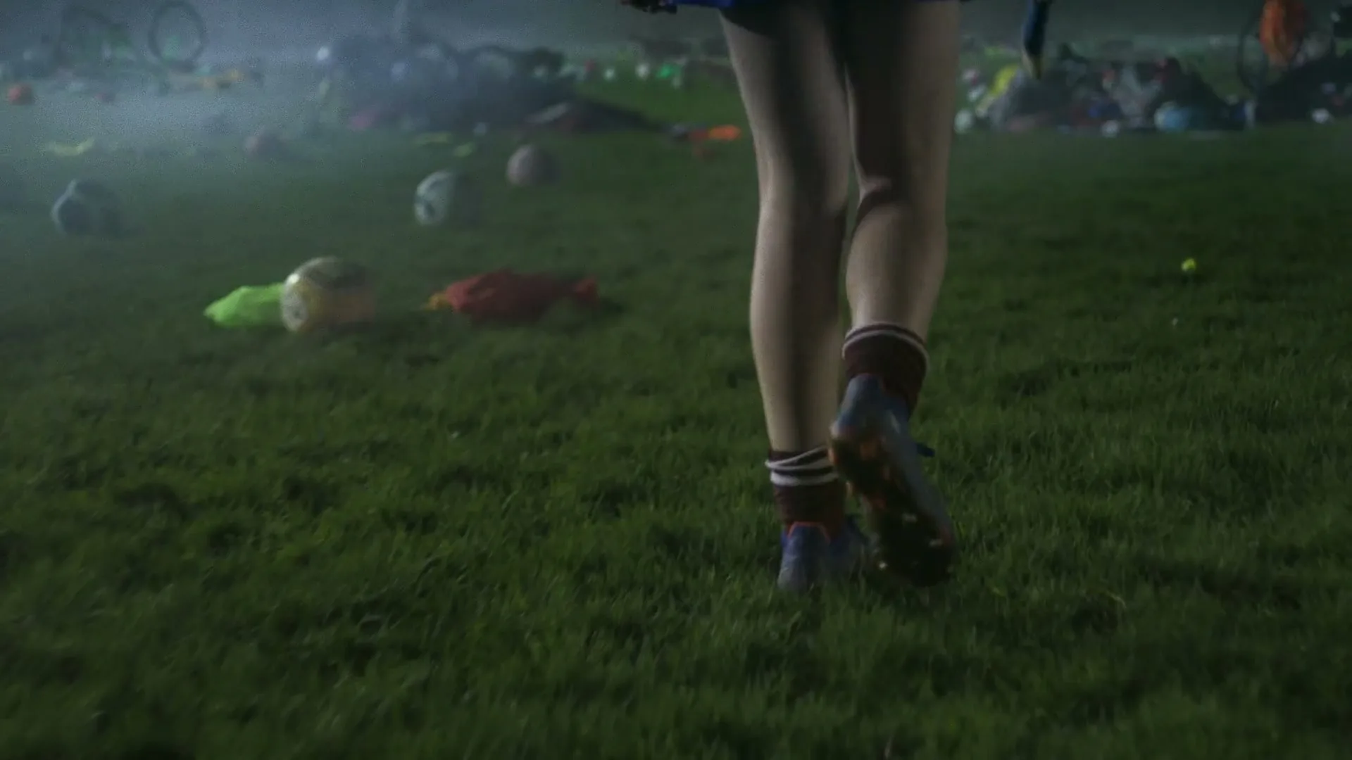 A young female athlete walks across a grassy field littered with sports equipment at night, her legs and feet in focus.