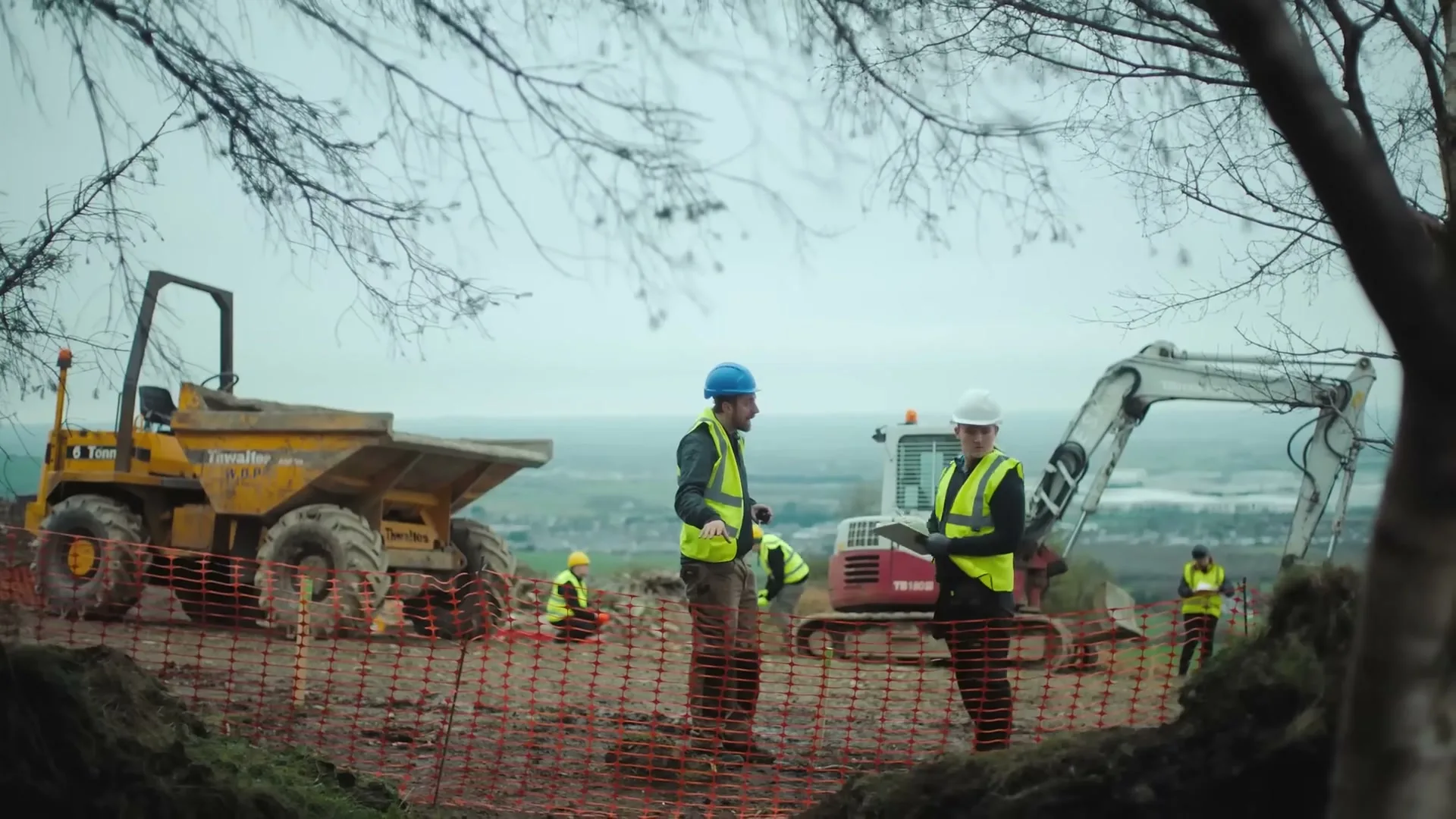 Two male construction workers in hard hats and high-vis vests discuss plans on a tablet at a muddy construction site with heavy machinery and other workers in the background.