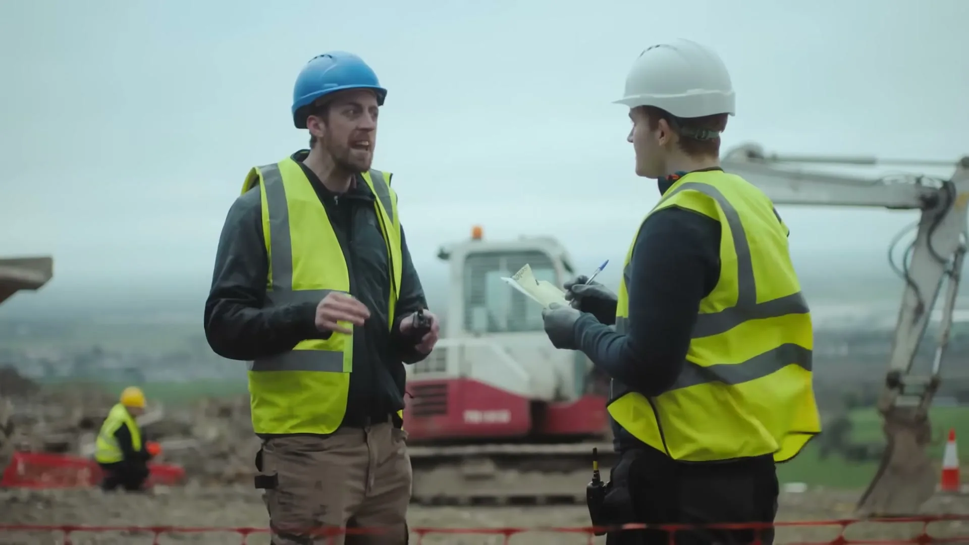 Two male construction workers in hard hats and high-vis vests discuss plans on a construction site with an excavator in the background.