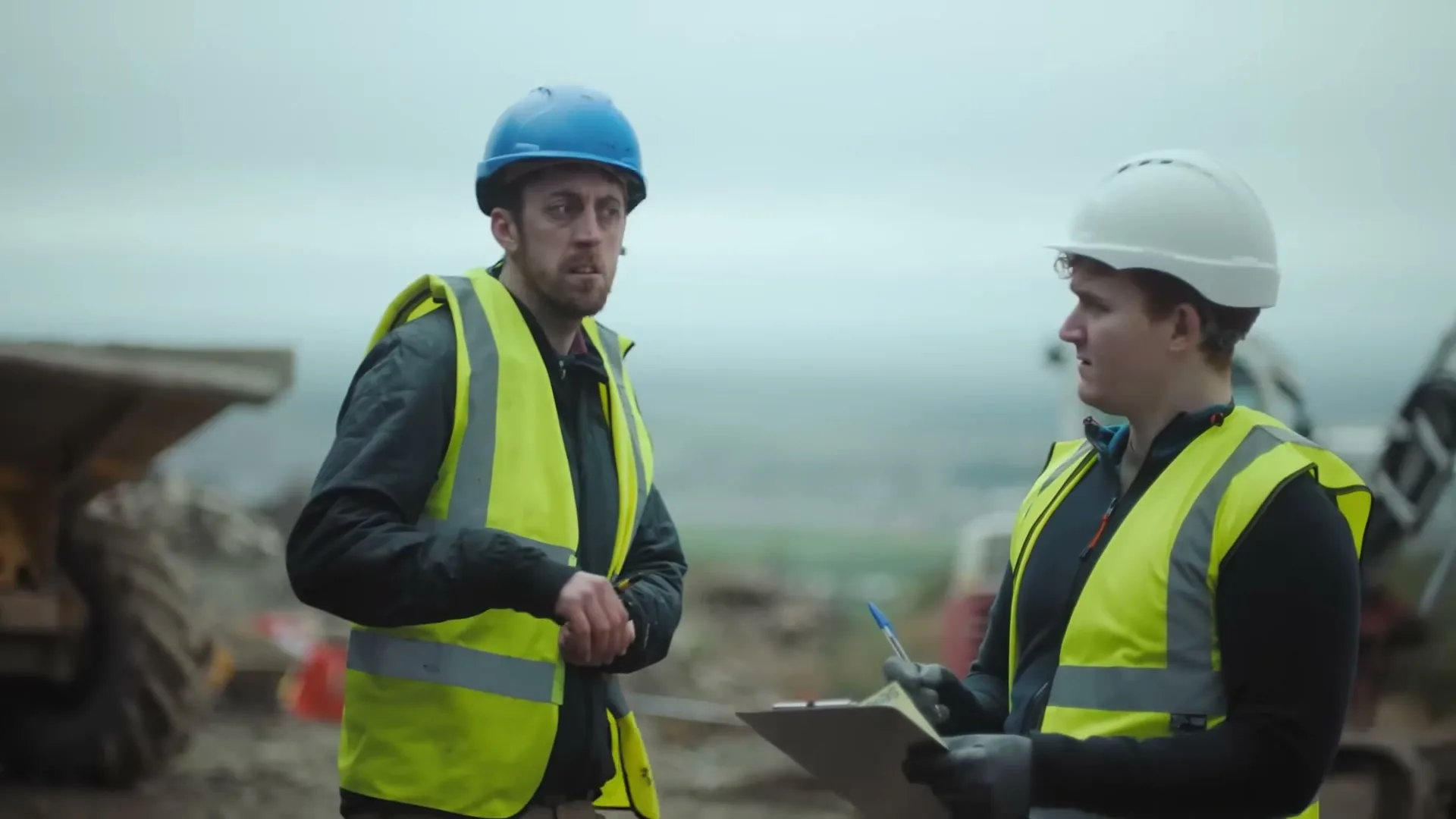 Two male construction workers in hard hats and high-vis vests stand on a muddy construction site, one holding a clipboard and pen.