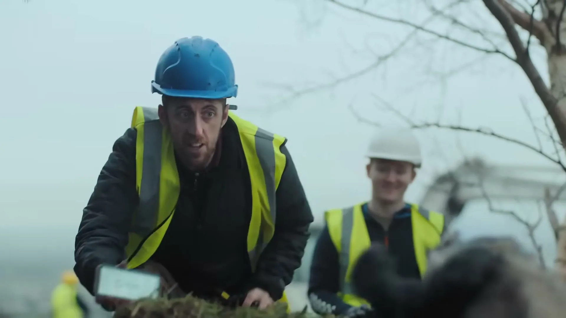 A male construction worker in a blue hard hat and high-vis jacket shows a smartphone screen to a badger, while another worker smiles in the background at a construction site.