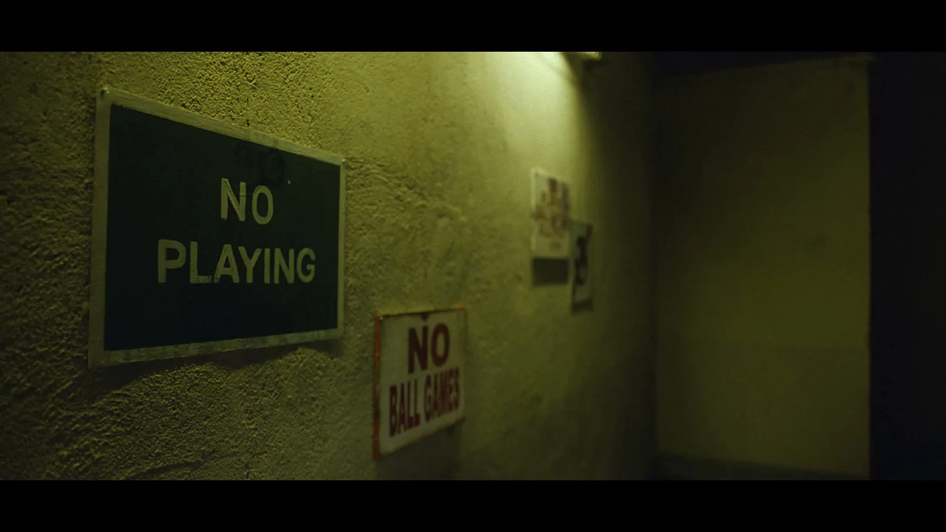 A dark, textured wall with two signs, one black with "NO PLAYING" in yellow, and another red with "NO BALL GAMES" in white, is dimly lit in an interior space.