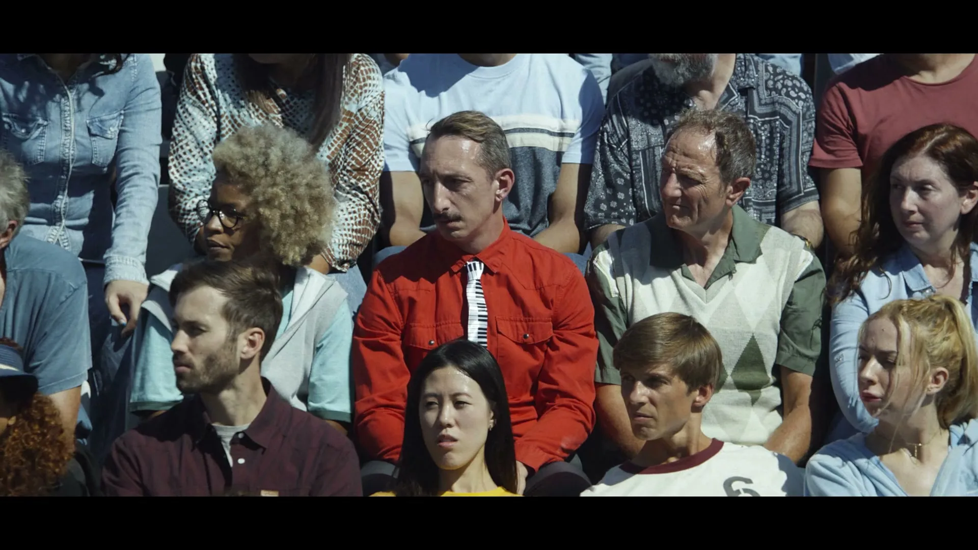 A diverse group of people, including a man in a red shirt and piano tie, sit in stadium bleachers, looking bored and unimpressed.