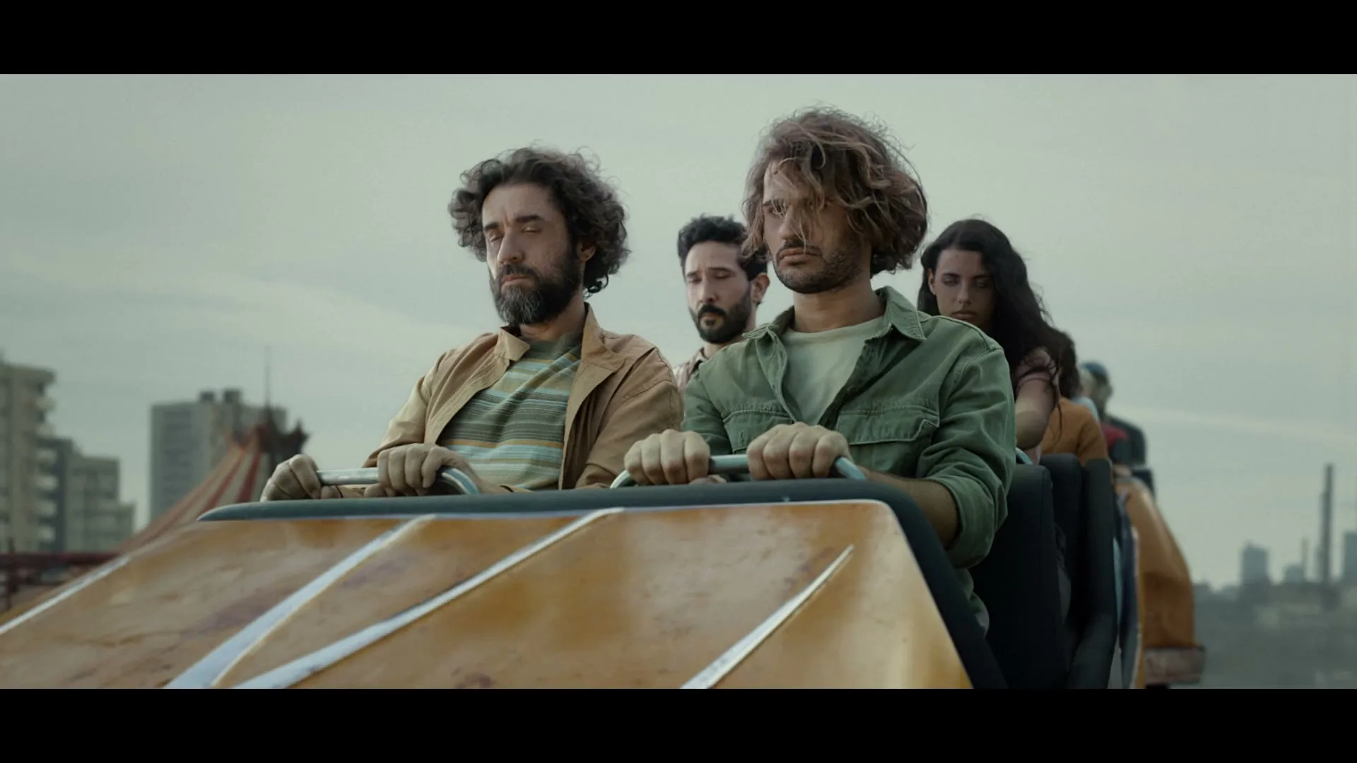 A group of four adults with curly hair ride a roller coaster with serious expressions, against a cloudy sky and urban background.