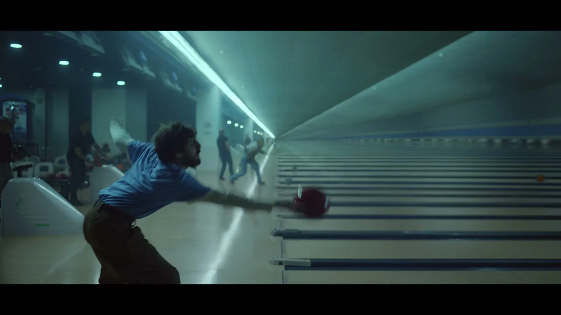 A man with a beard in a blue shirt bowls a red ball down a long alley in a brightly lit bowling alley.