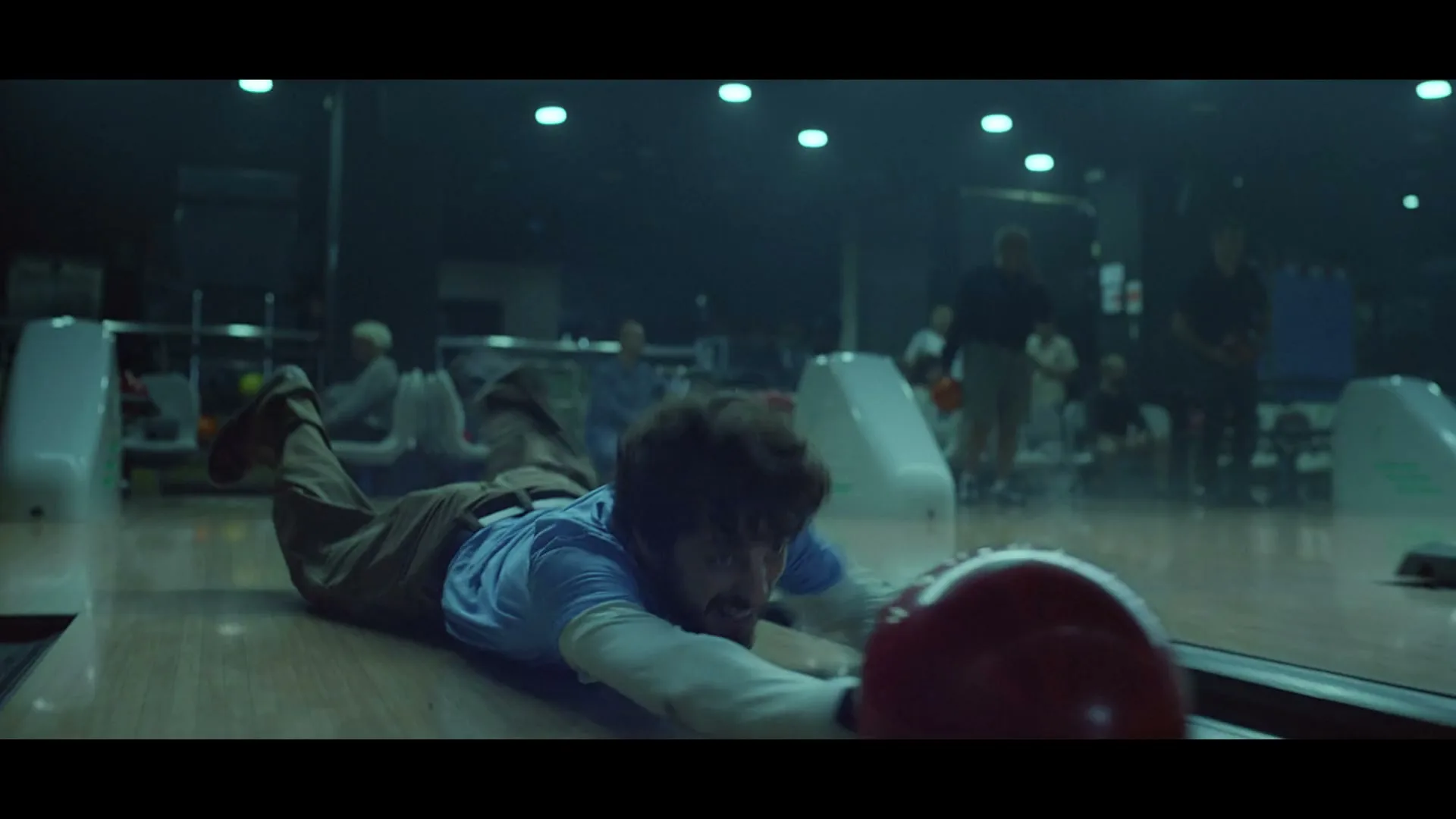A man with a beard, wearing a blue shirt and brown pants, slides on the bowling alley floor, reaching for a red bowling ball.