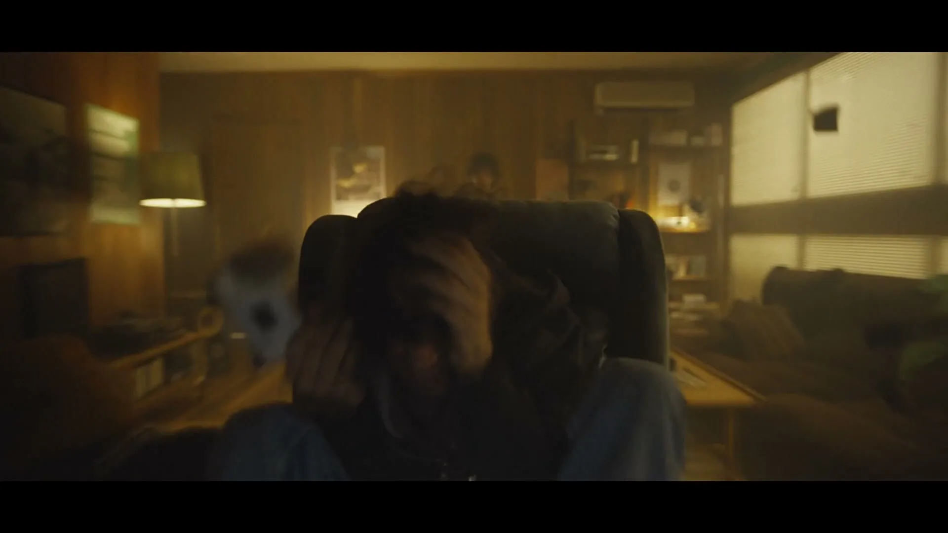 A young man with long hair, wearing a hoodie, screams in terror while holding a white game controller in a dimly lit room, as the camera rapidly pulls back.
