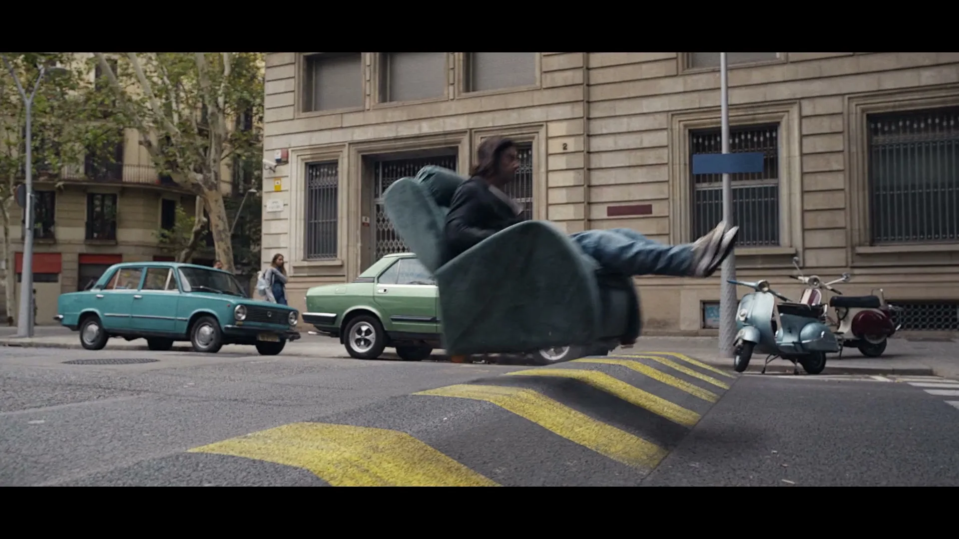 A man with long hair and a beard sits in a green armchair, floating over a speed bump on a city street with cars and buildings.