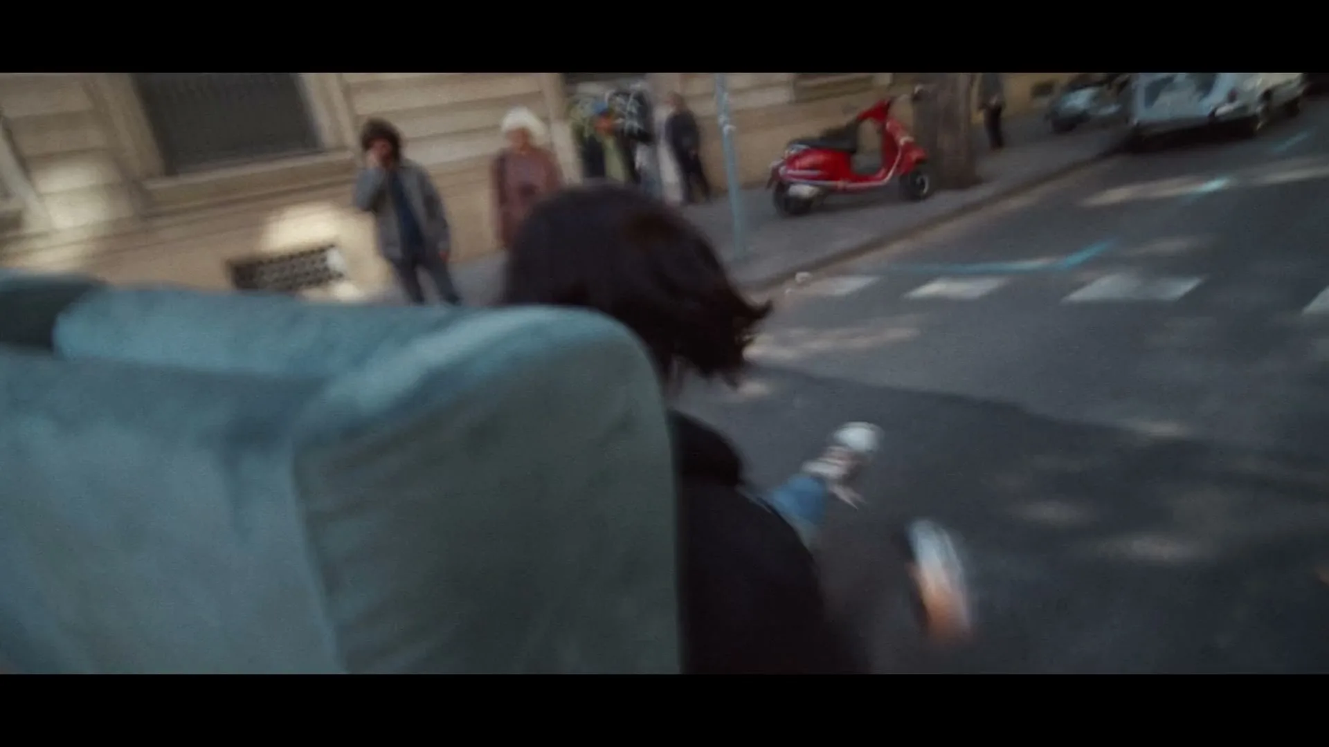 A person with dark hair, wearing a black top and jeans, is seen from behind, sitting on a teal sofa that is sliding down a city street.