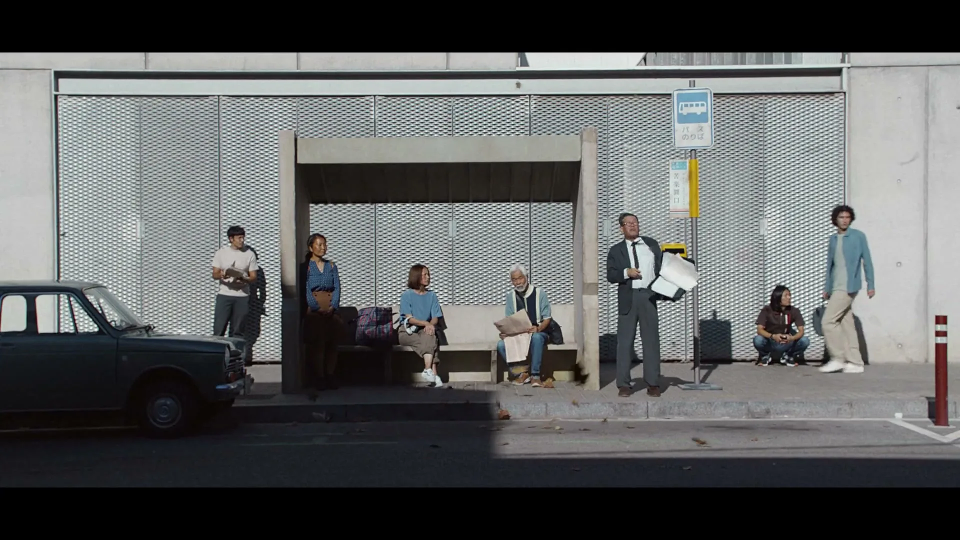 A diverse group of people wait at a bus stop, looking bored, until a man in a suit throws papers into the air, surprising everyone.