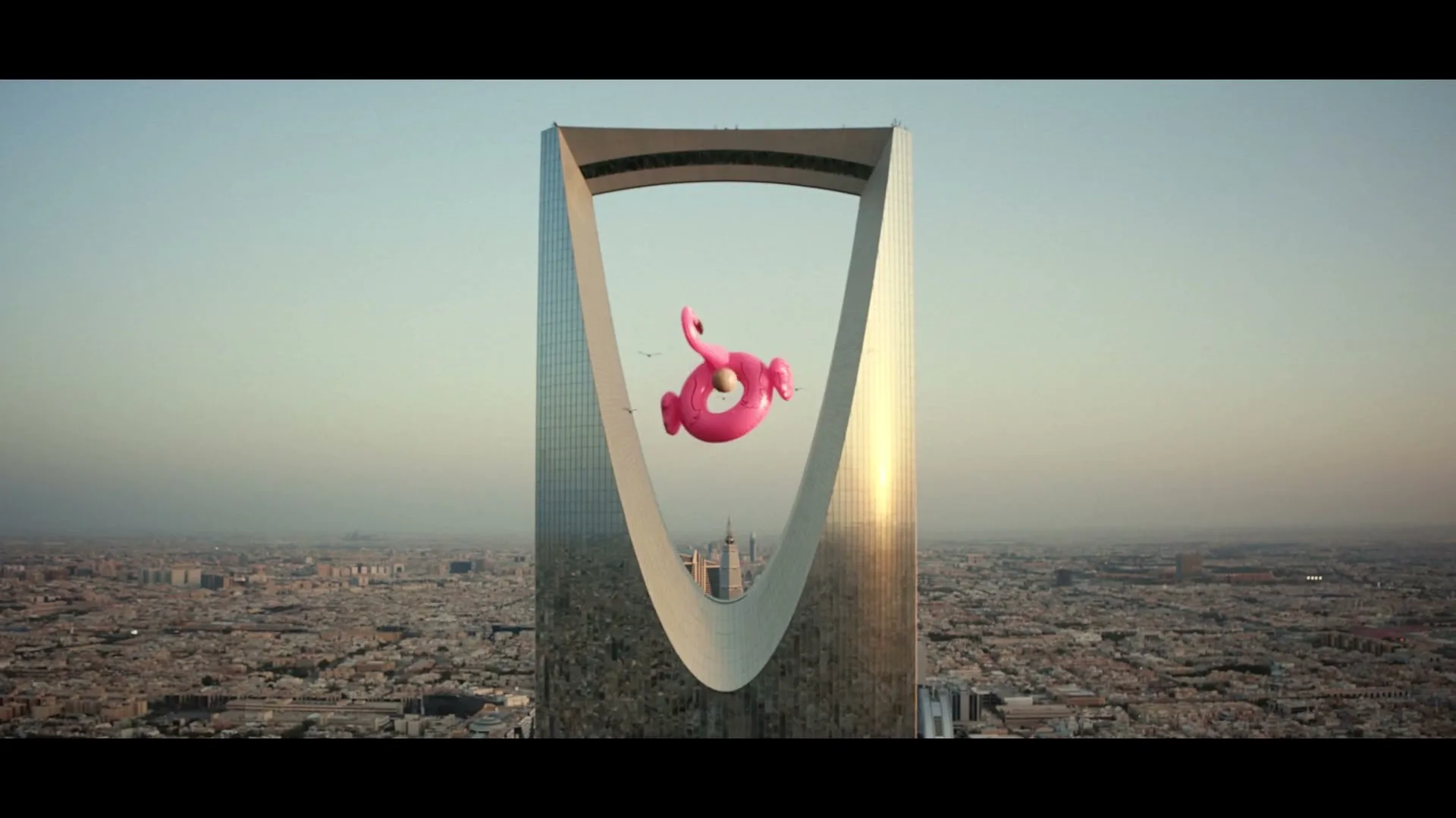 A giant pink flamingo inflatable floats in front of the Kingdom Centre Tower in Riyadh, Saudi Arabia, with the city skyline visible below.