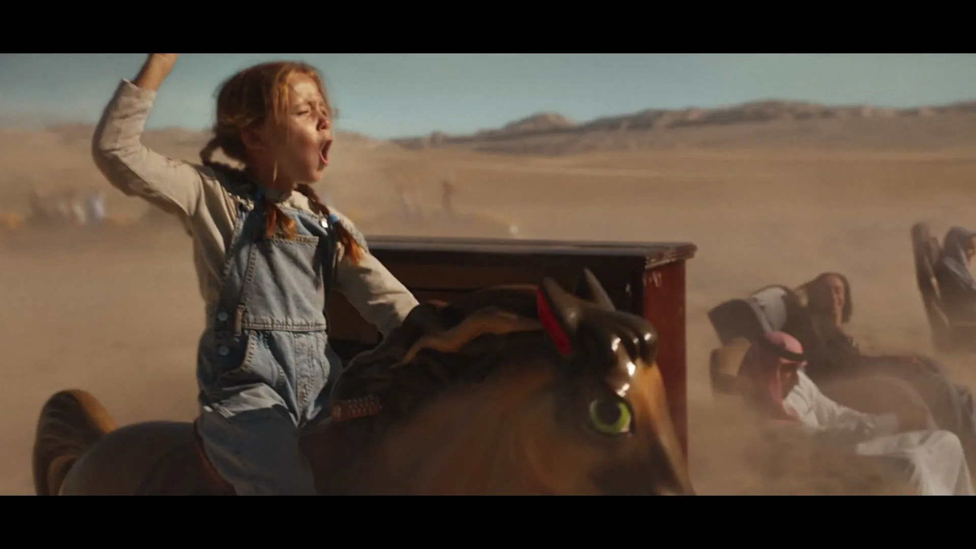 A young girl with pigtails, wearing denim overalls, rides a mechanical horse in a dusty desert landscape, shouting with excitement.