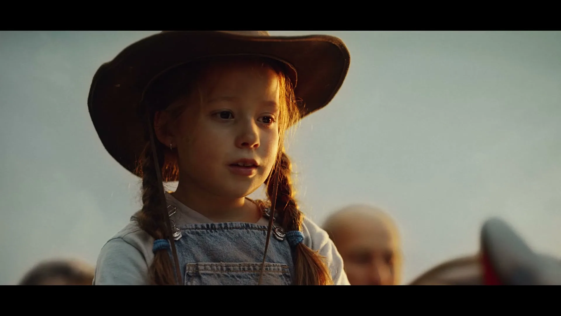 A young girl with pigtails and a cowboy hat looks on with a serious expression, with blurred figures in the background.