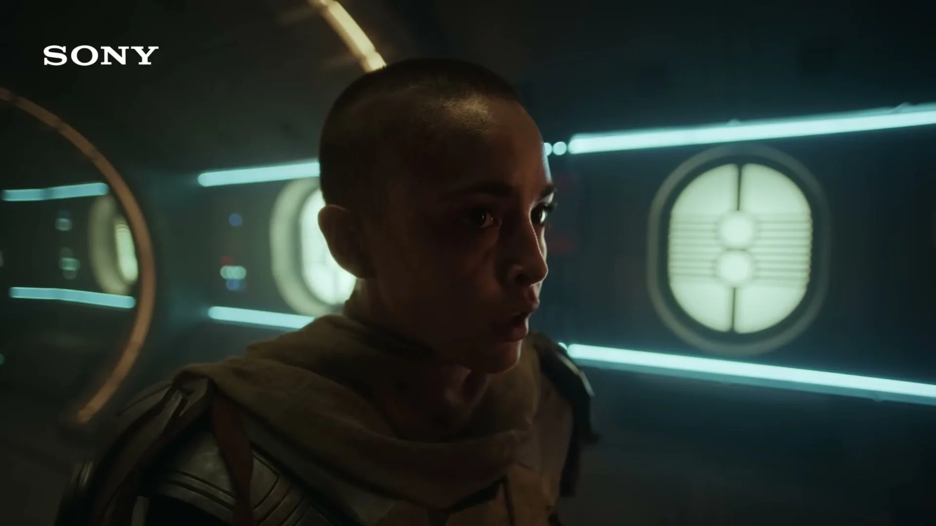 A woman with a shaved head and a determined expression looks around a futuristic, dimly lit spaceship interior, wearing a brown scarf and armor.