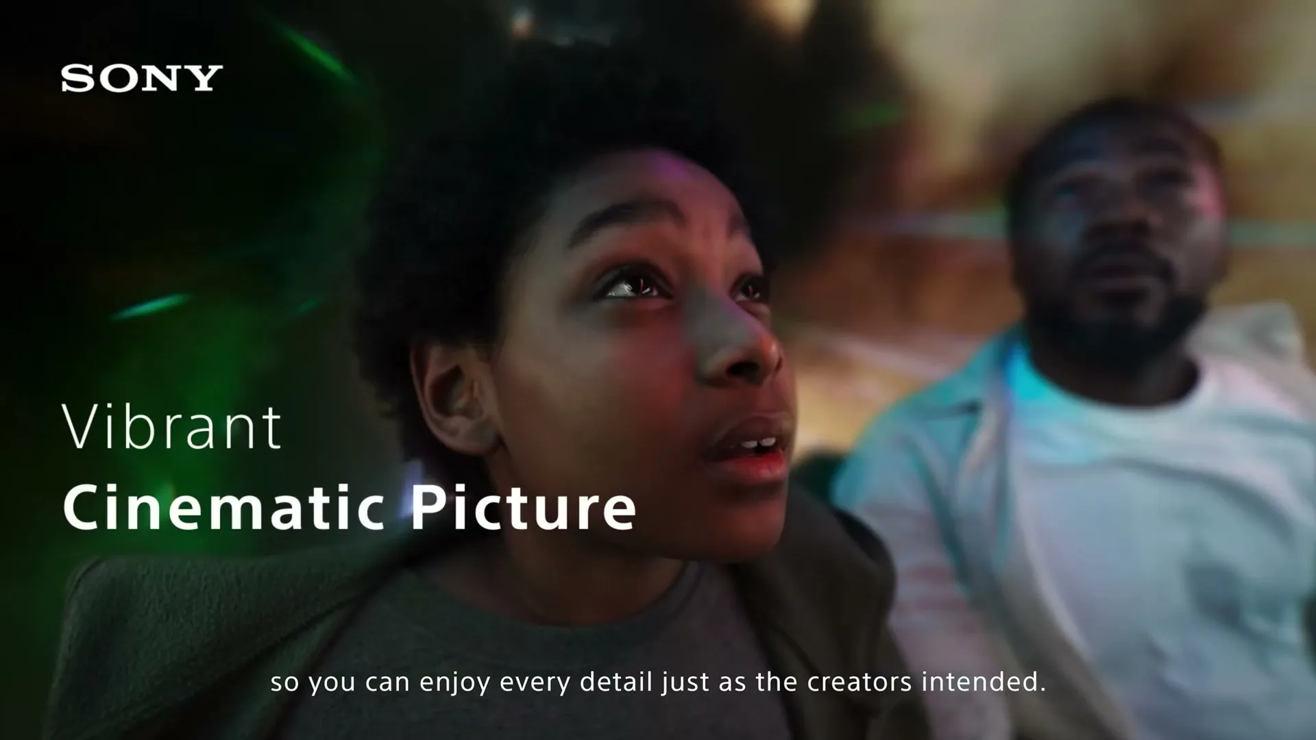 A young boy and a man watch a screen with vibrant colors reflecting on their faces, in a dark room.