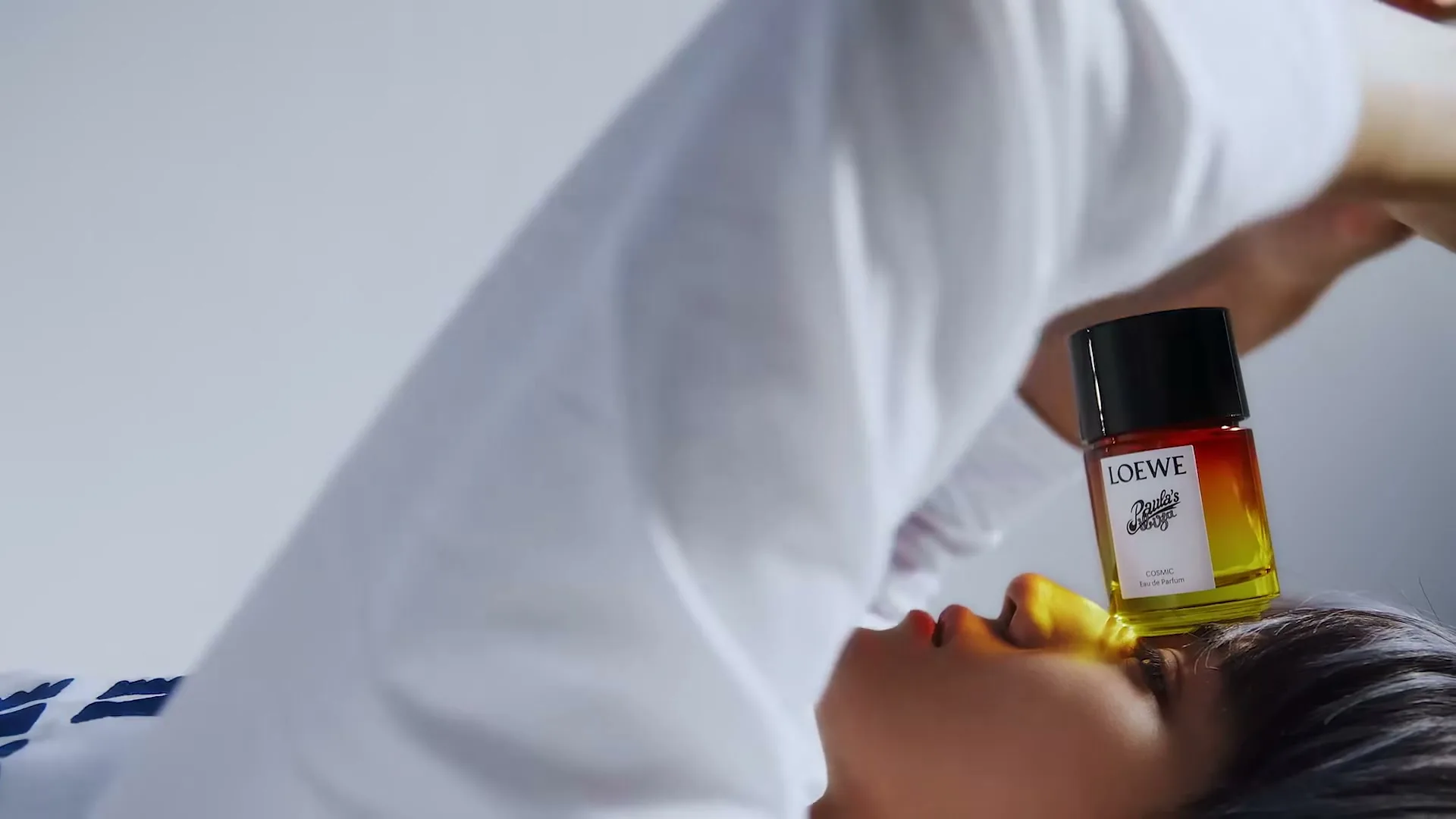A person with dark hair and a white shirt lies on their back with a Loewe perfume bottle on their forehead, in a bright, minimalist setting.