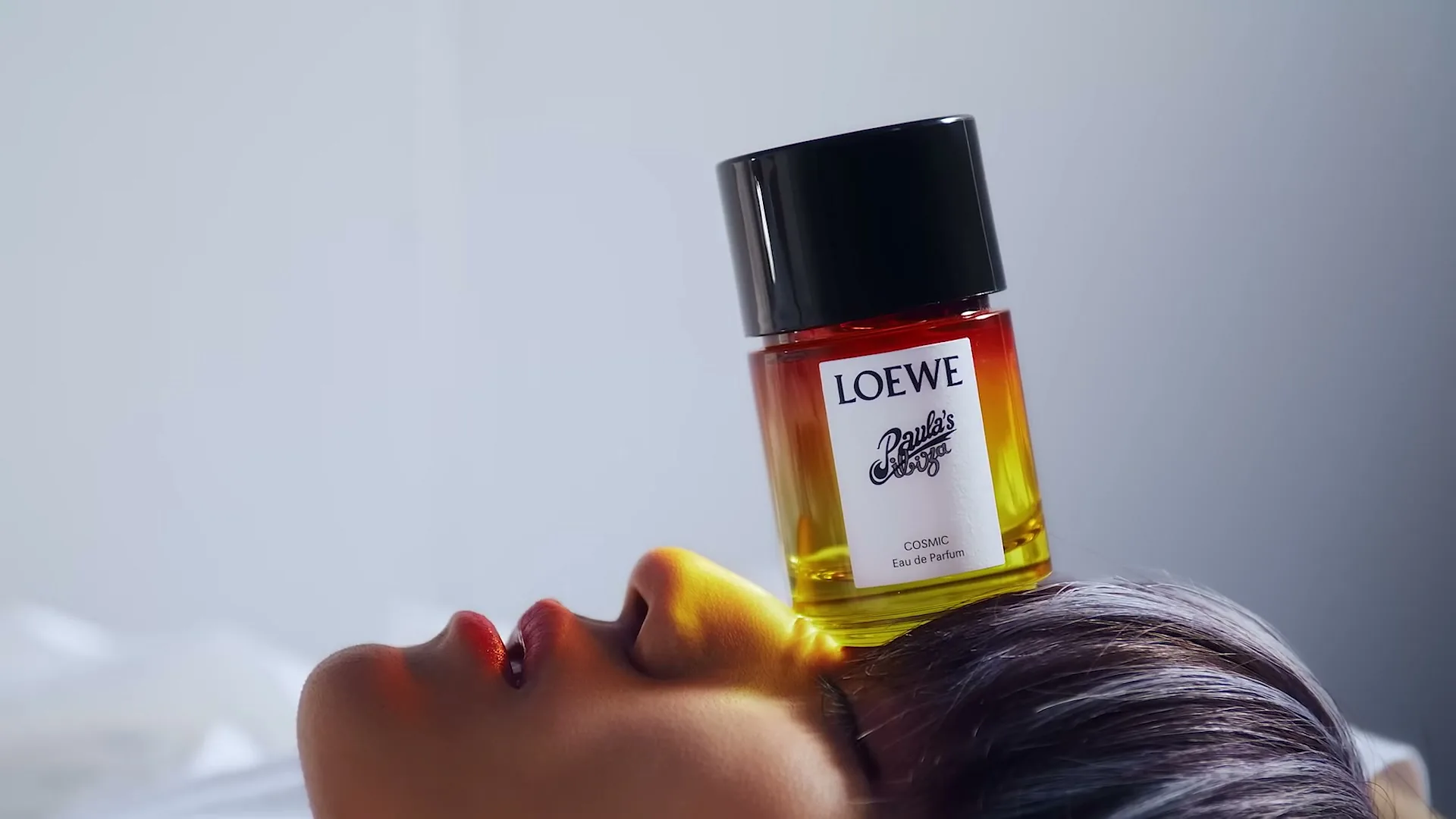 A perfume bottle with a gradient of red, orange, and yellow sits on the forehead of a person with red lipstick, looking up.