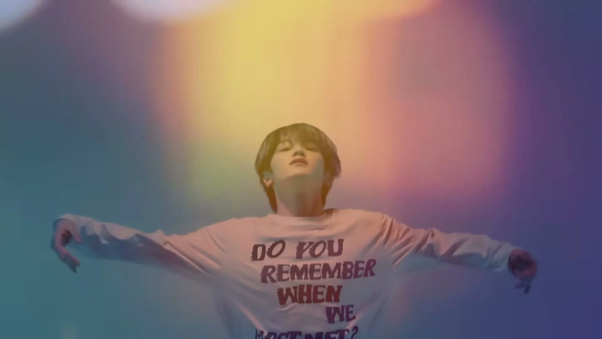 A young person with dark hair and a white long-sleeved shirt with text on it floats in a colorful, ethereal space with arms outstretched.