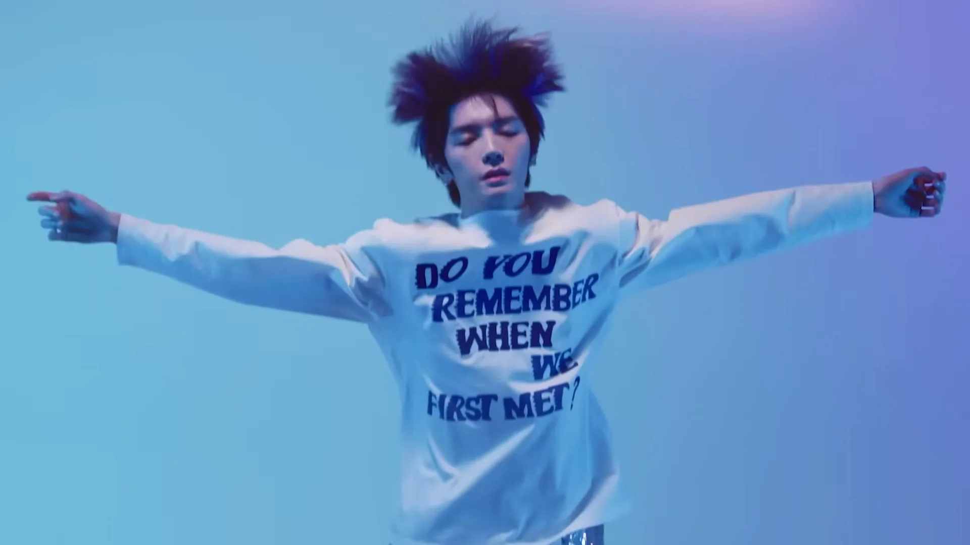 A young male with spiky hair and a white sweatshirt with text jumps with arms outstretched against a blue and purple background.