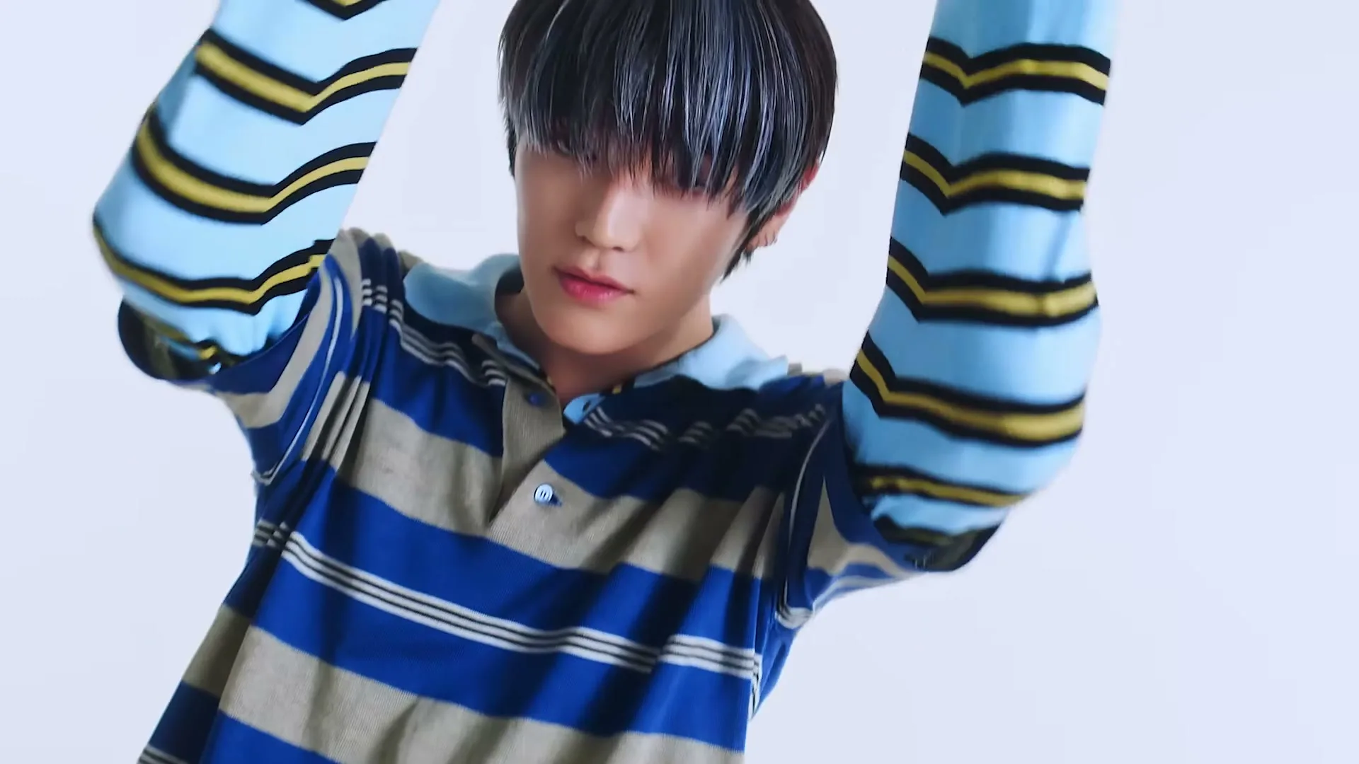 A young man with dark hair and a striped polo shirt poses with his arms raised, looking directly at the camera in a bright studio.