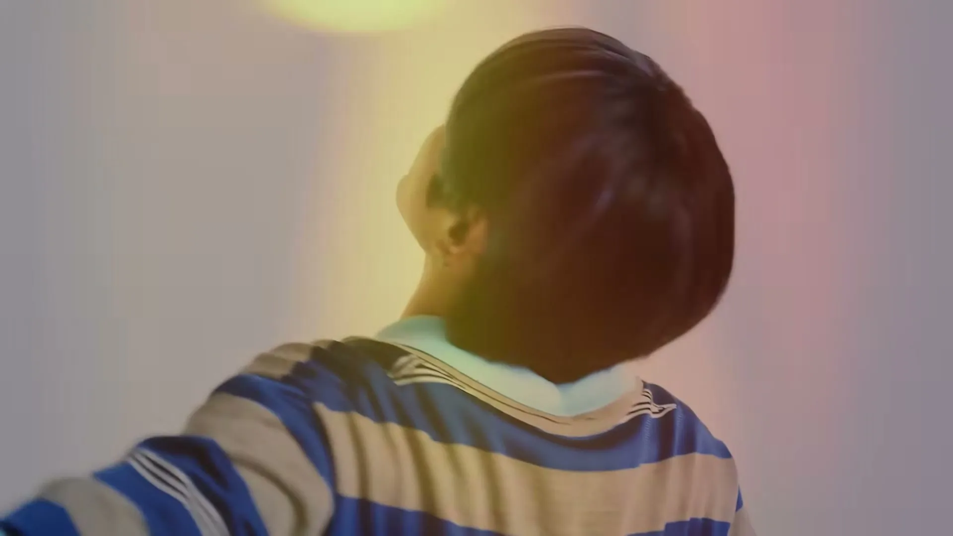 A young man with dark hair and a striped shirt spins around in a bright, white studio, then lies on a white sheet with a hand near his head, looking at the camera.