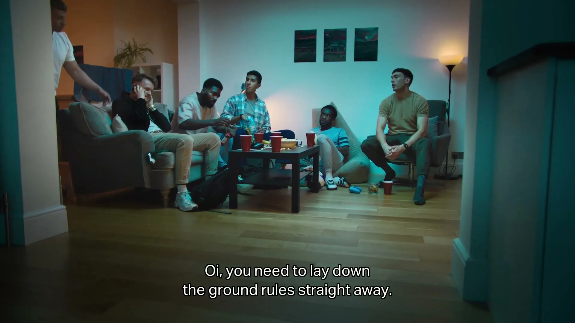 A diverse group of young men are gathered in a living room, some sitting on a sofa and others on the floor, watching something intently.
