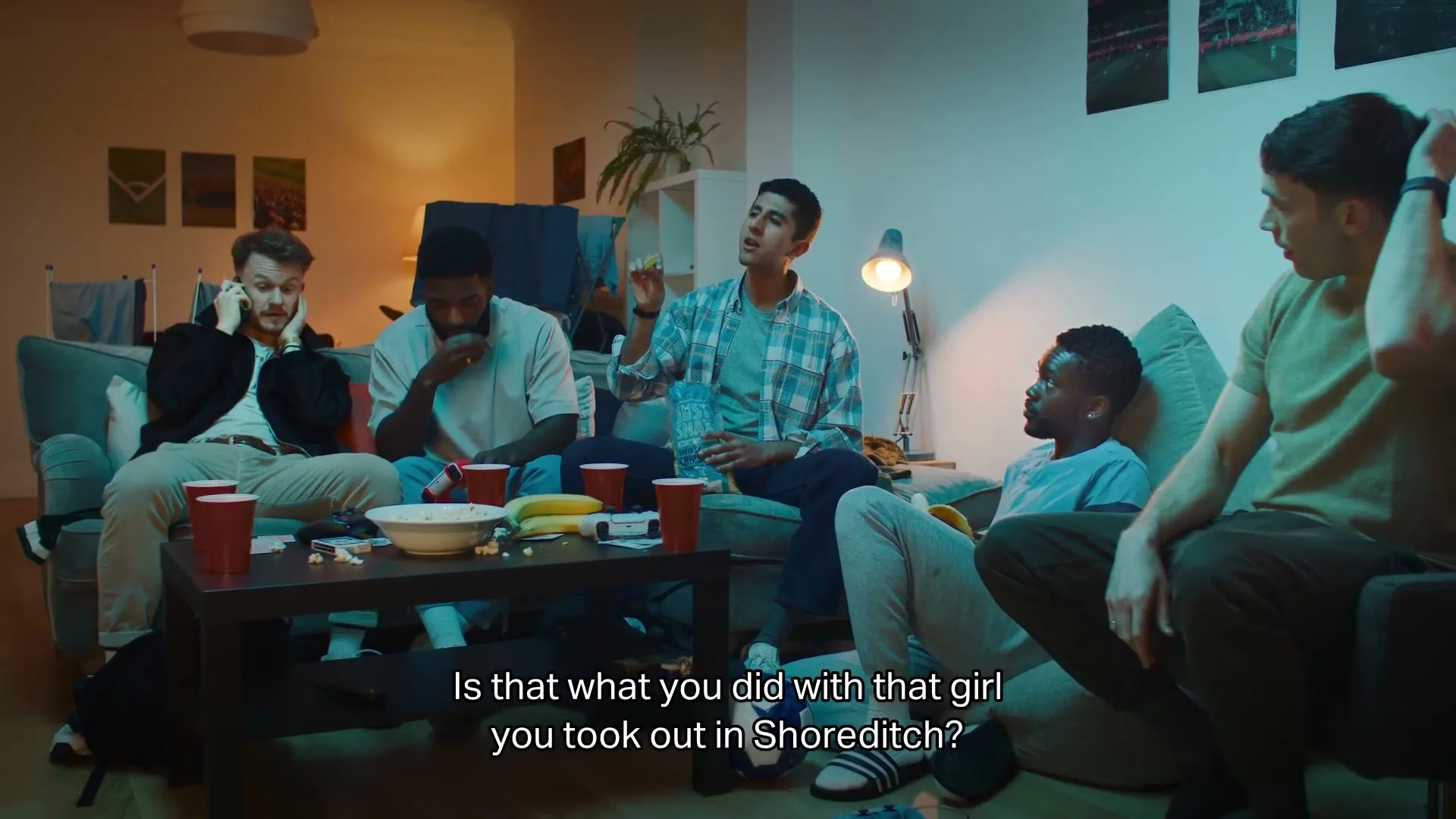 Five young men are hanging out in a dimly lit living room, talking and relaxing on couches and beanbags, with snacks and drinks on a coffee table.