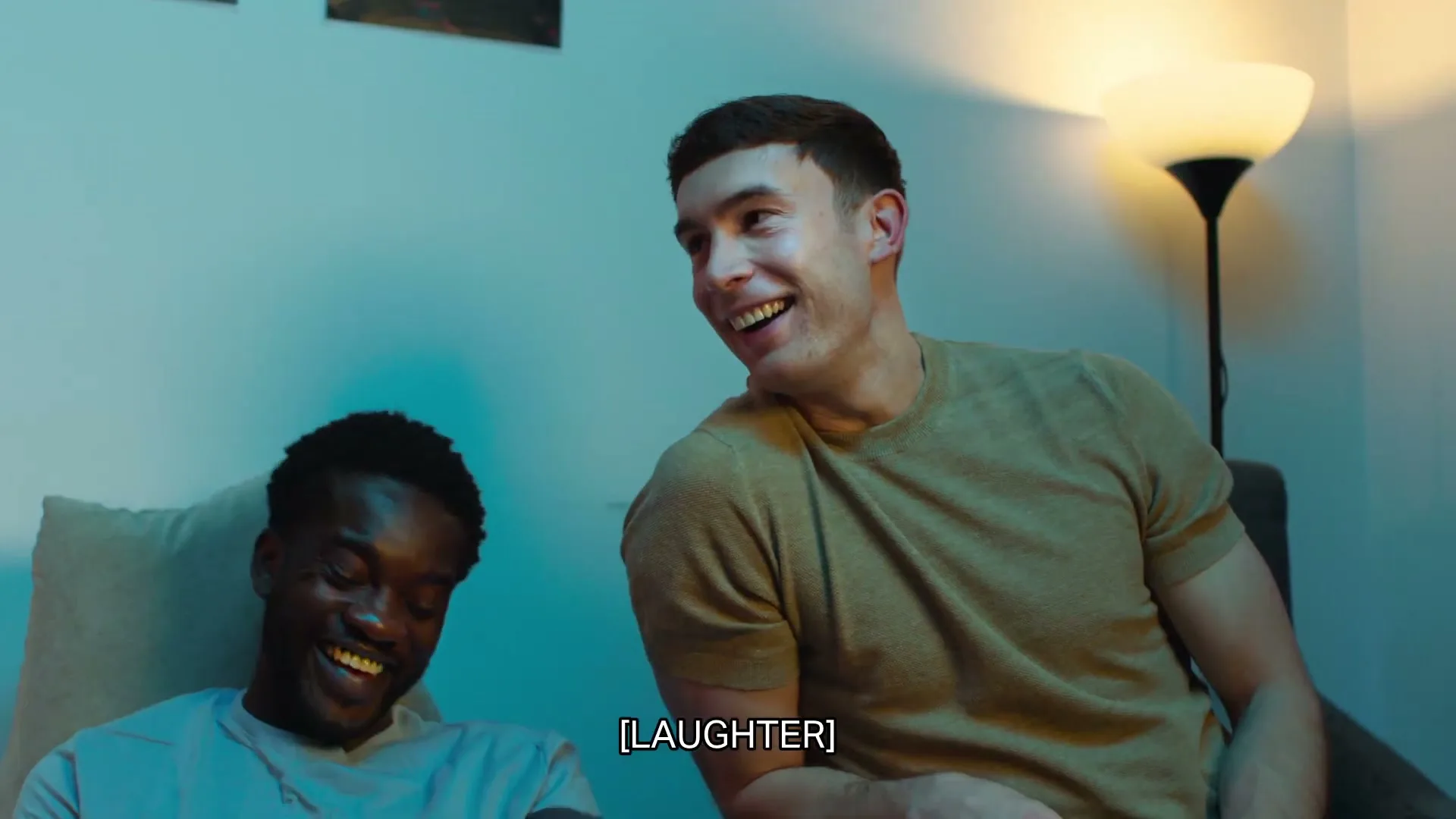 Two young men, one Black and one White, sit on a sofa in a living room at night, laughing and looking to the right.