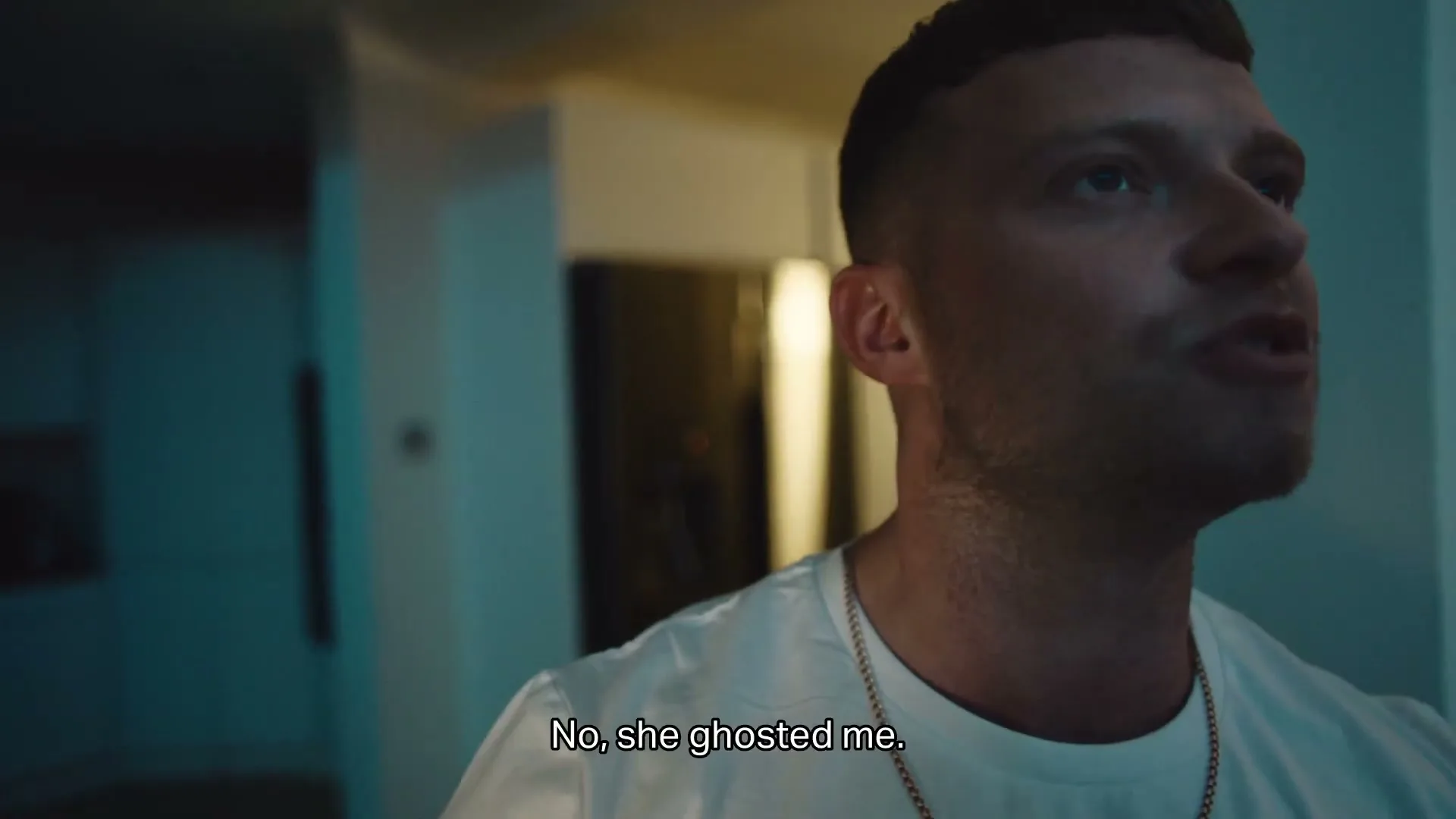 A young man with a short haircut and white t-shirt looks around and speaks in a dimly lit interior.
