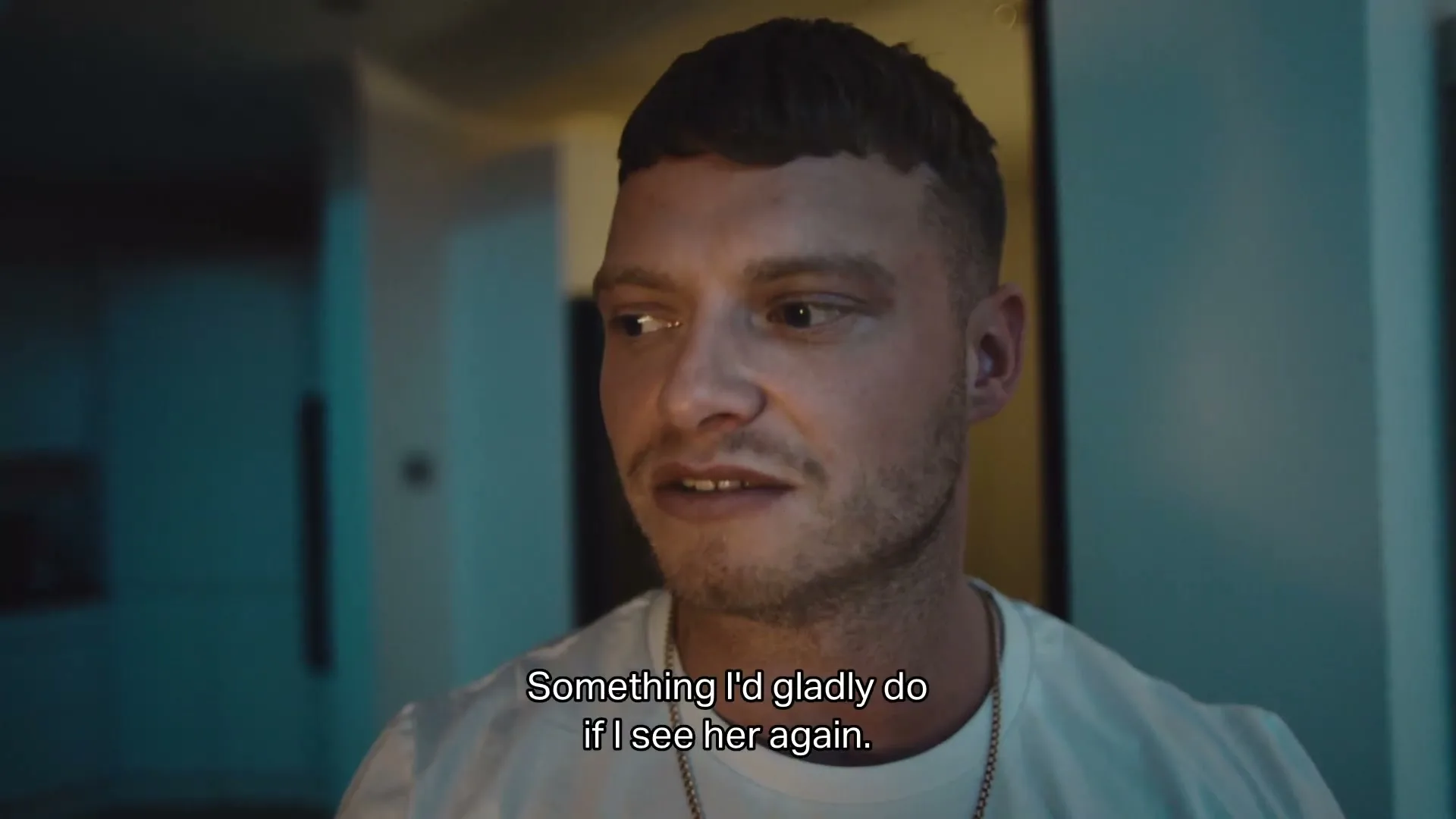 A young man with a short haircut and a white t-shirt looks around nervously in a dimly lit interior.