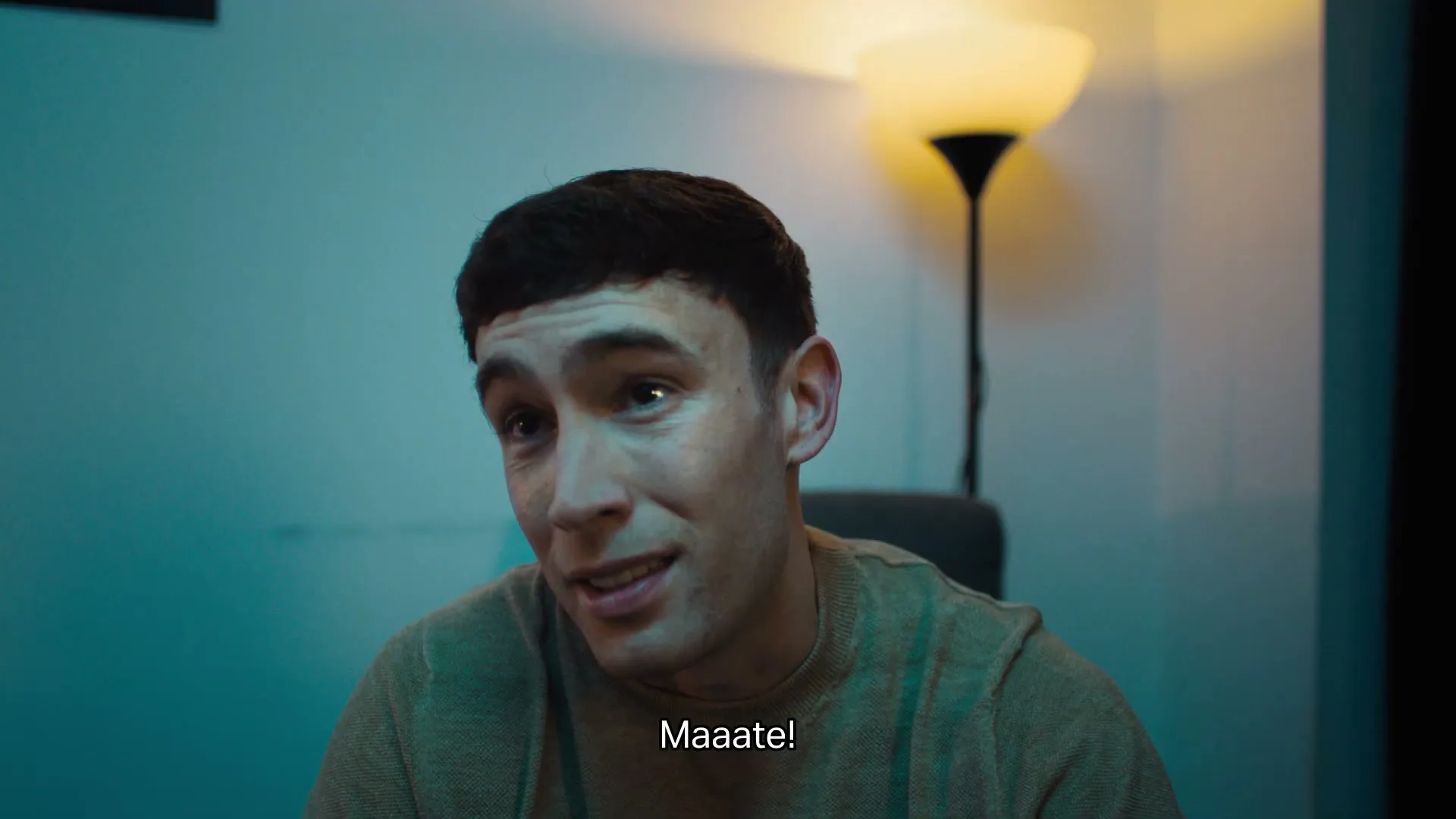 A young man with short dark hair, wearing a beige sweater, looks up and to the right, speaking in a dimly lit room.