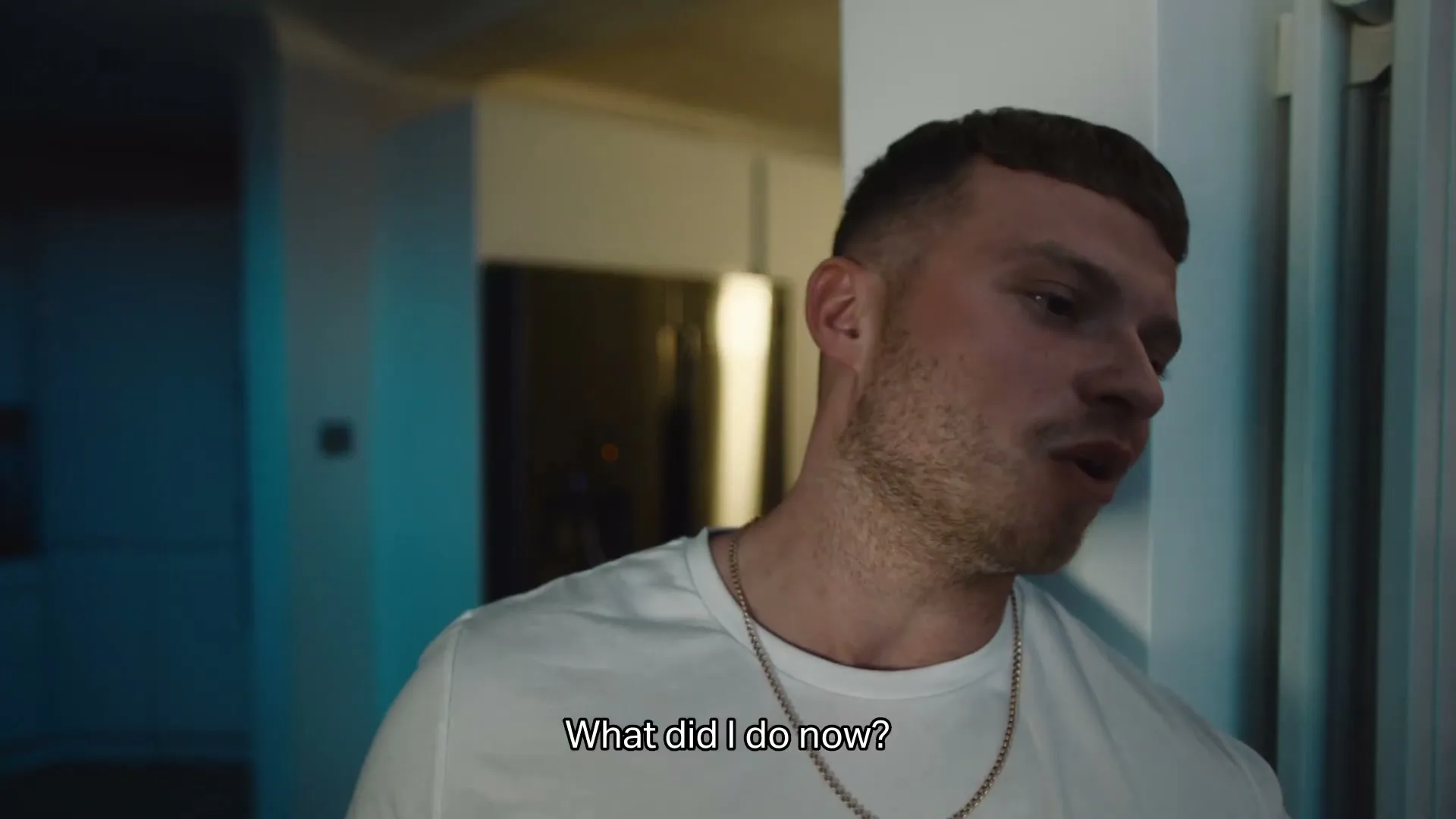 A young man with short hair and a white t-shirt leans against a wall, looking frustrated and speaking in an interior setting.