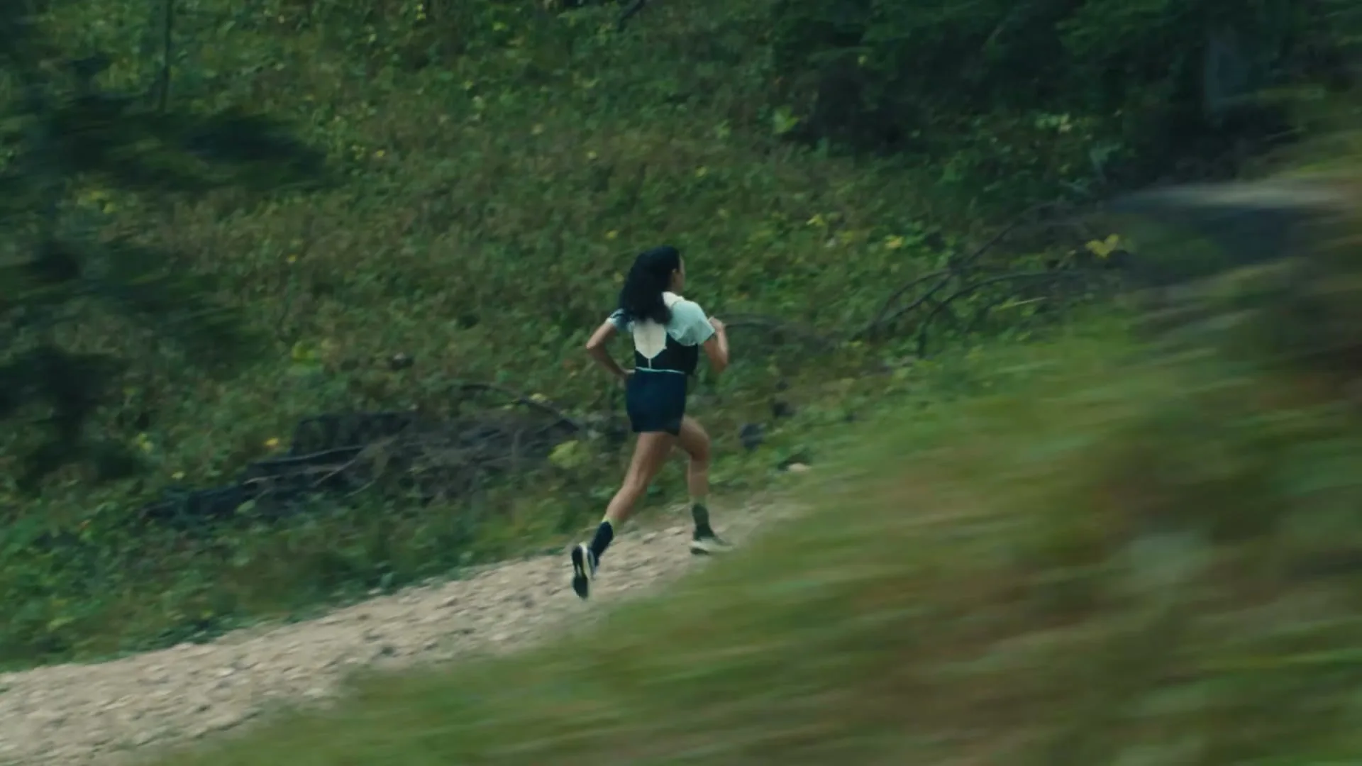 A woman with dark hair, wearing a light blue top and dark shorts, runs uphill on a dirt path in a lush green forest.