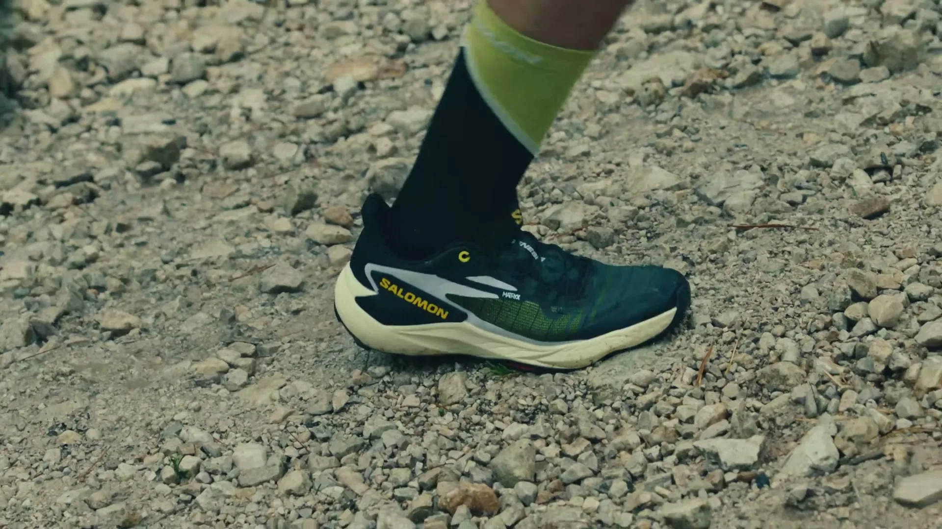 A person's foot wearing a Salomon running shoe steps onto a rocky, dirt path in an outdoor setting during the day.