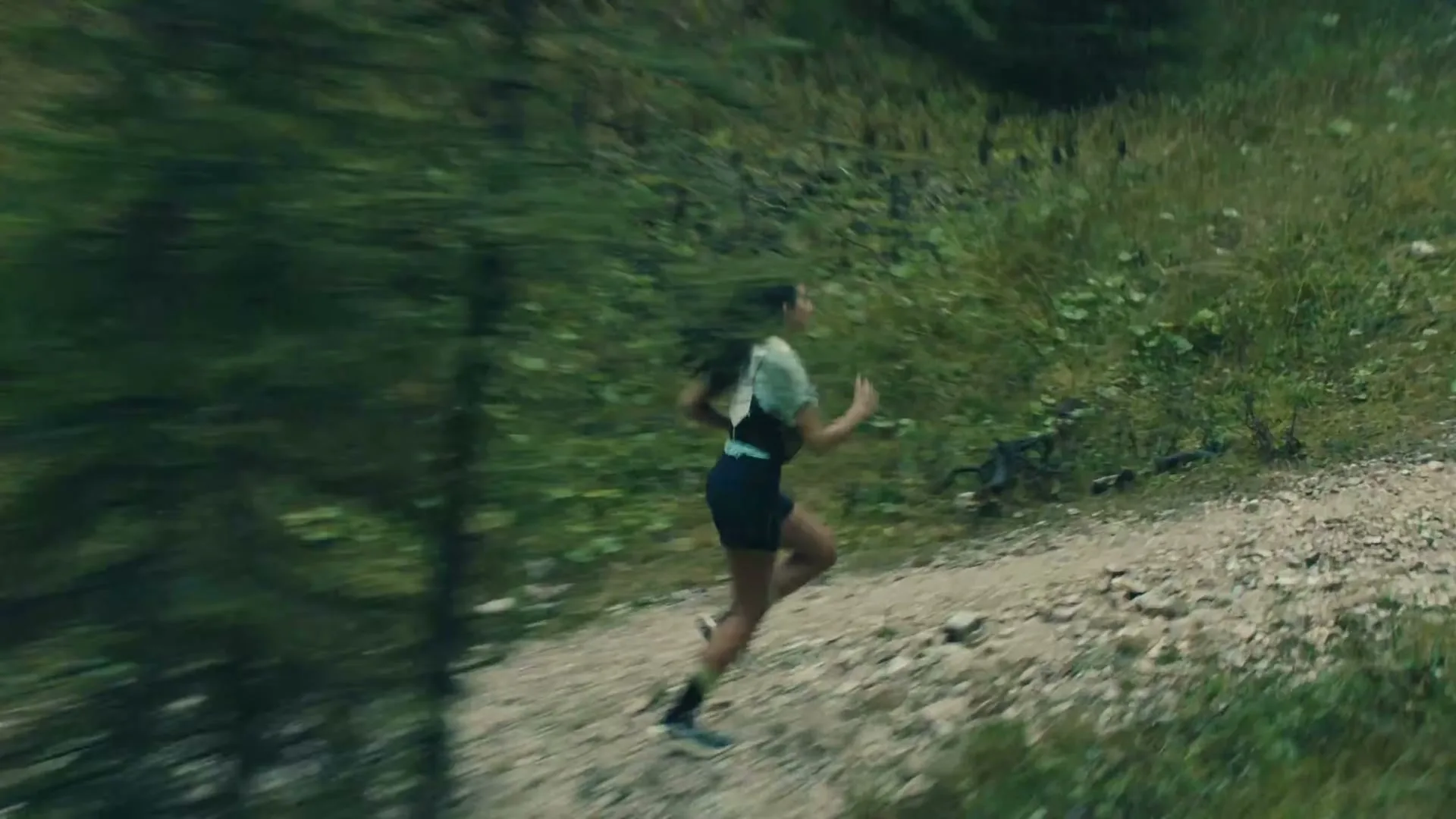A young woman in athletic wear runs uphill on a dirt path through a lush green forest on a sunny day.