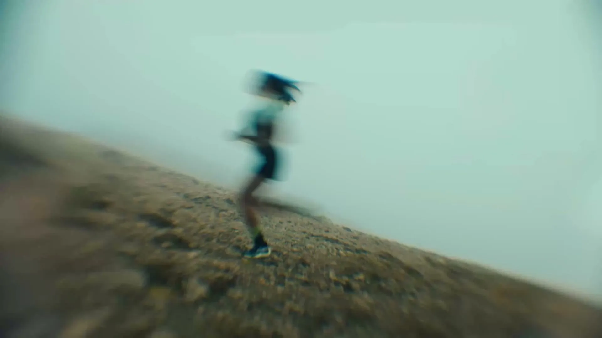 A blurry shot of a female runner in athletic wear, running uphill on a dirt path against a foggy, light blue sky.