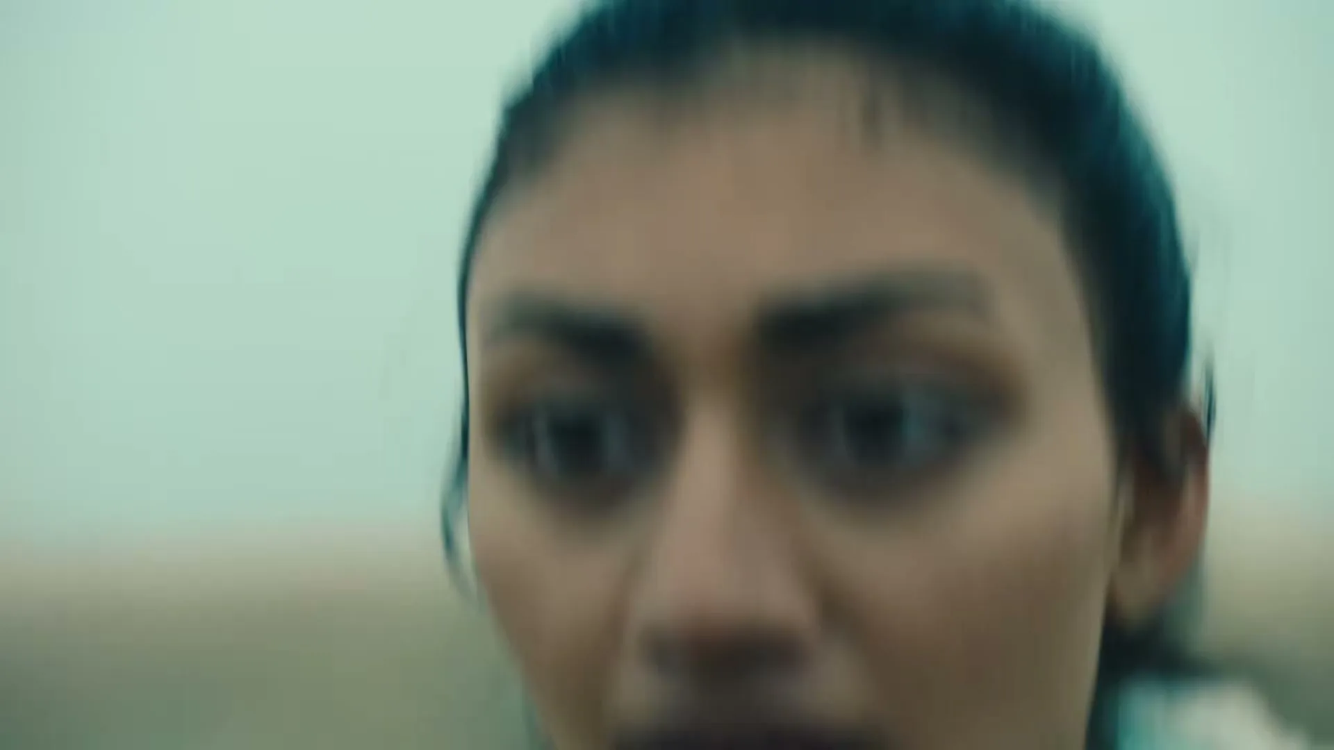 A woman with dark hair and a light blue shirt is running and looking around with a determined expression in an outdoor setting.