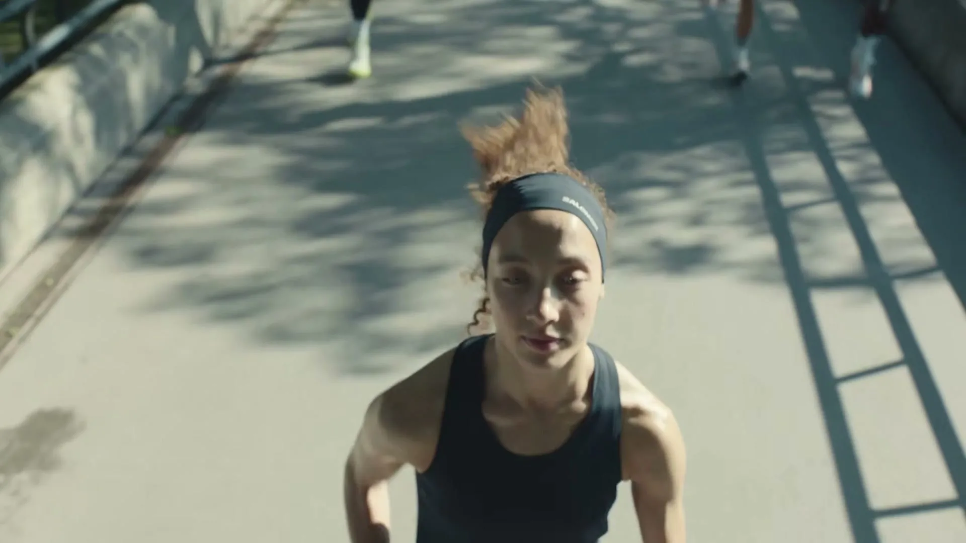 A female runner in a black tank top and headband runs towards the camera on a bridge, with other runners in the background on a sunny day.