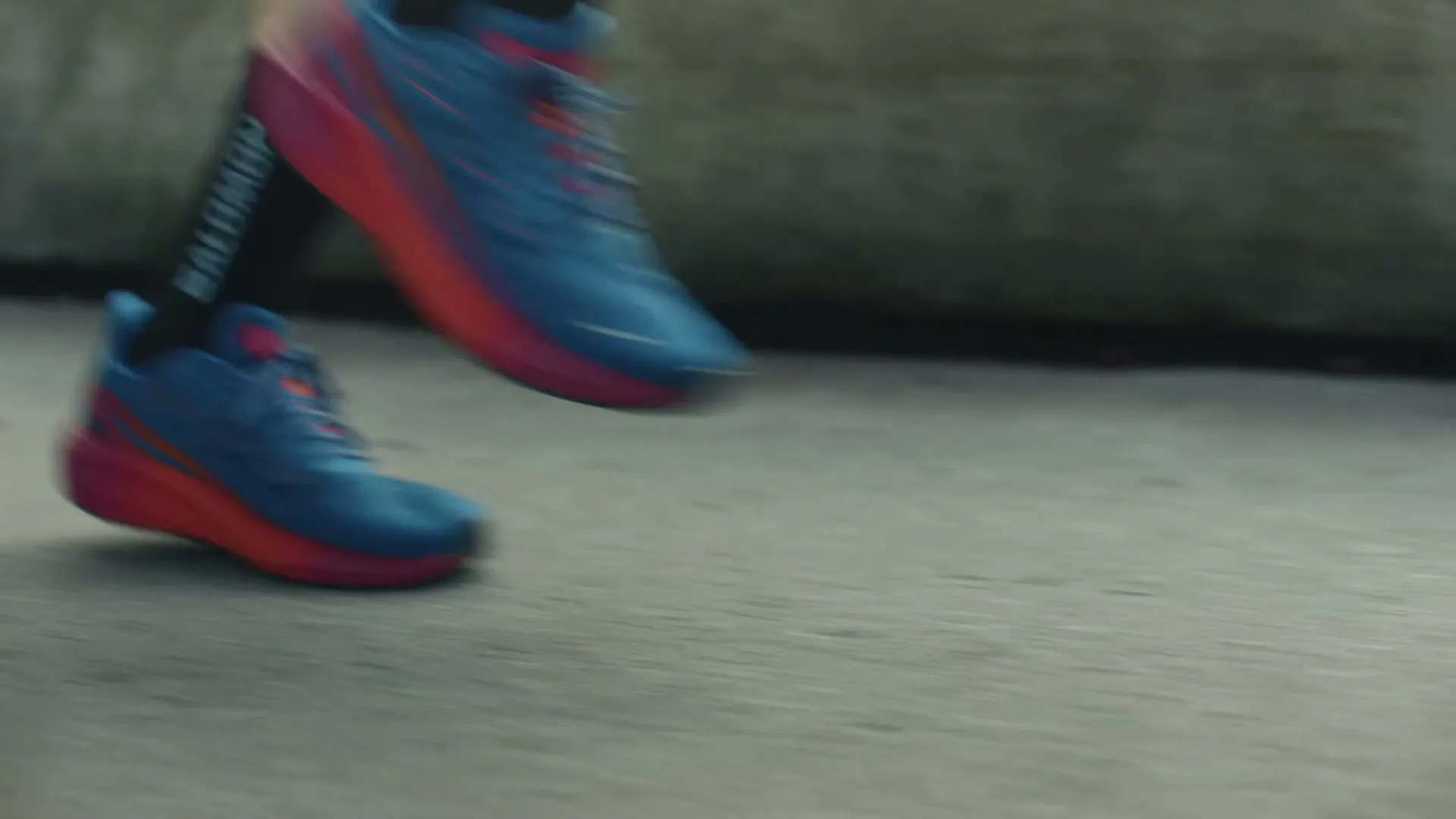 A person's feet wearing blue and red running shoes and black socks with the Salomon logo are seen running on a paved surface.