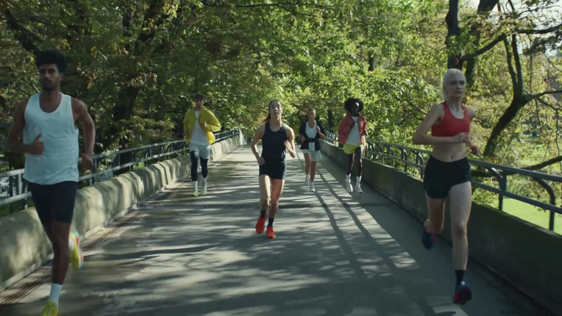 A diverse group of six runners in athletic wear runs across a bridge in a park on a sunny day.