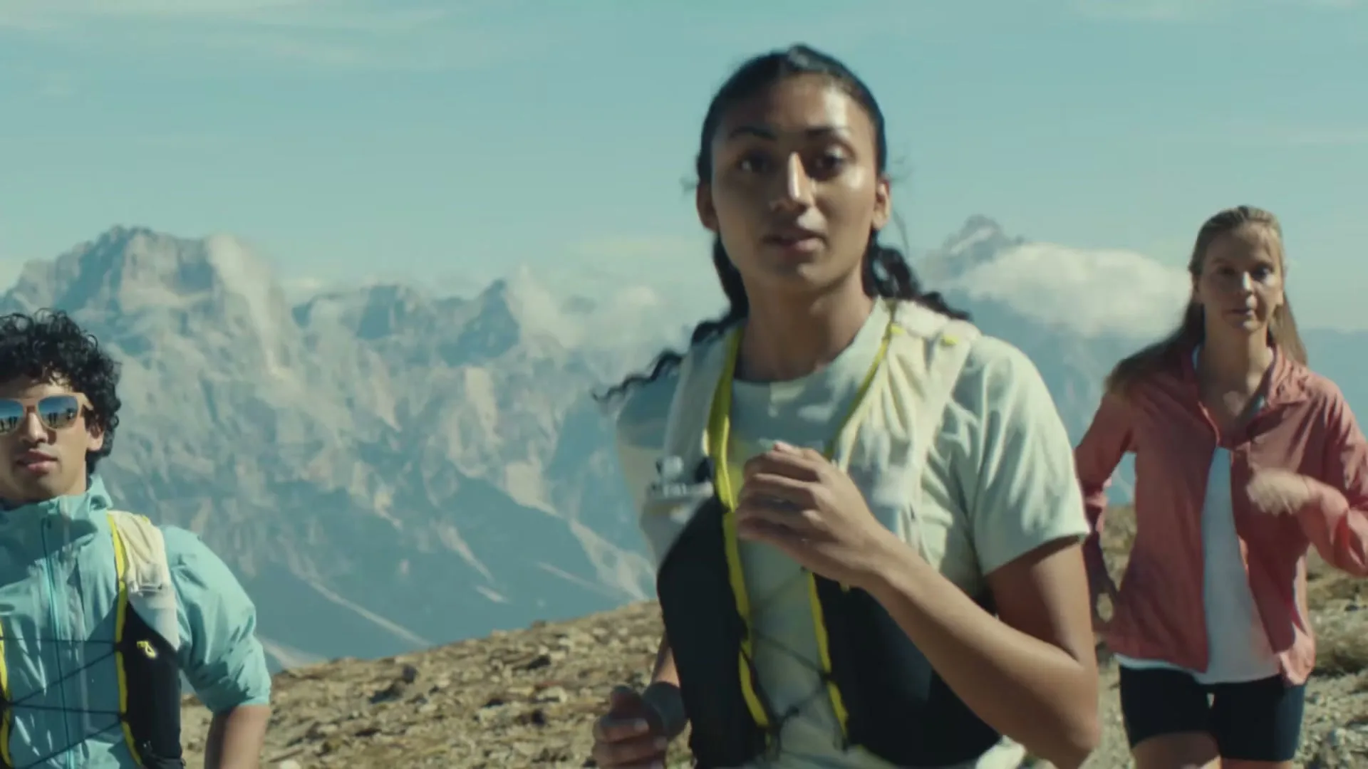 Three young adults, two women and one man, are trail running uphill on a rocky mountain path under a clear sky.