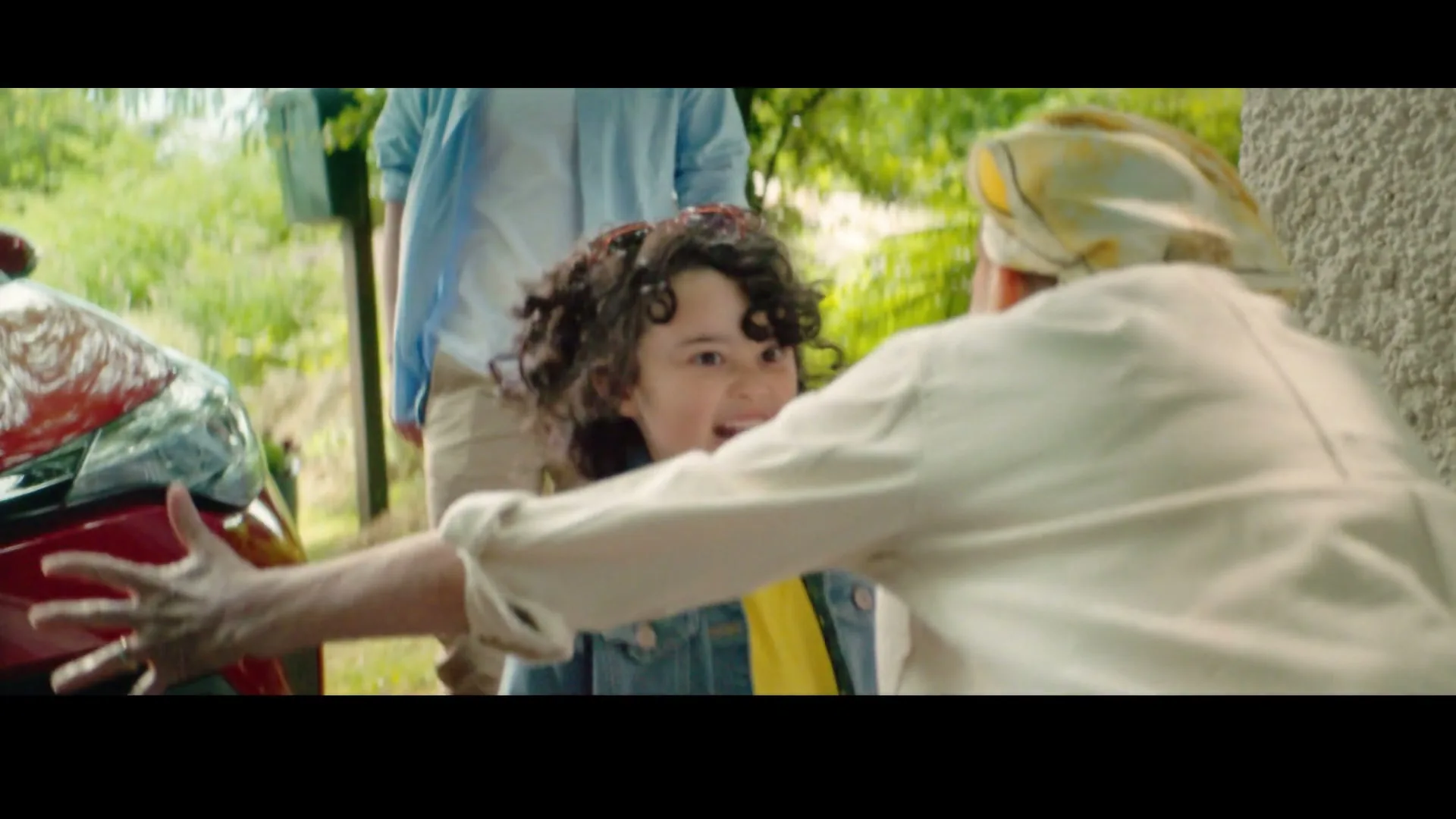 A young girl with curly hair and sunglasses on her head runs to hug an older woman, while a man stands in the background next to a red car.