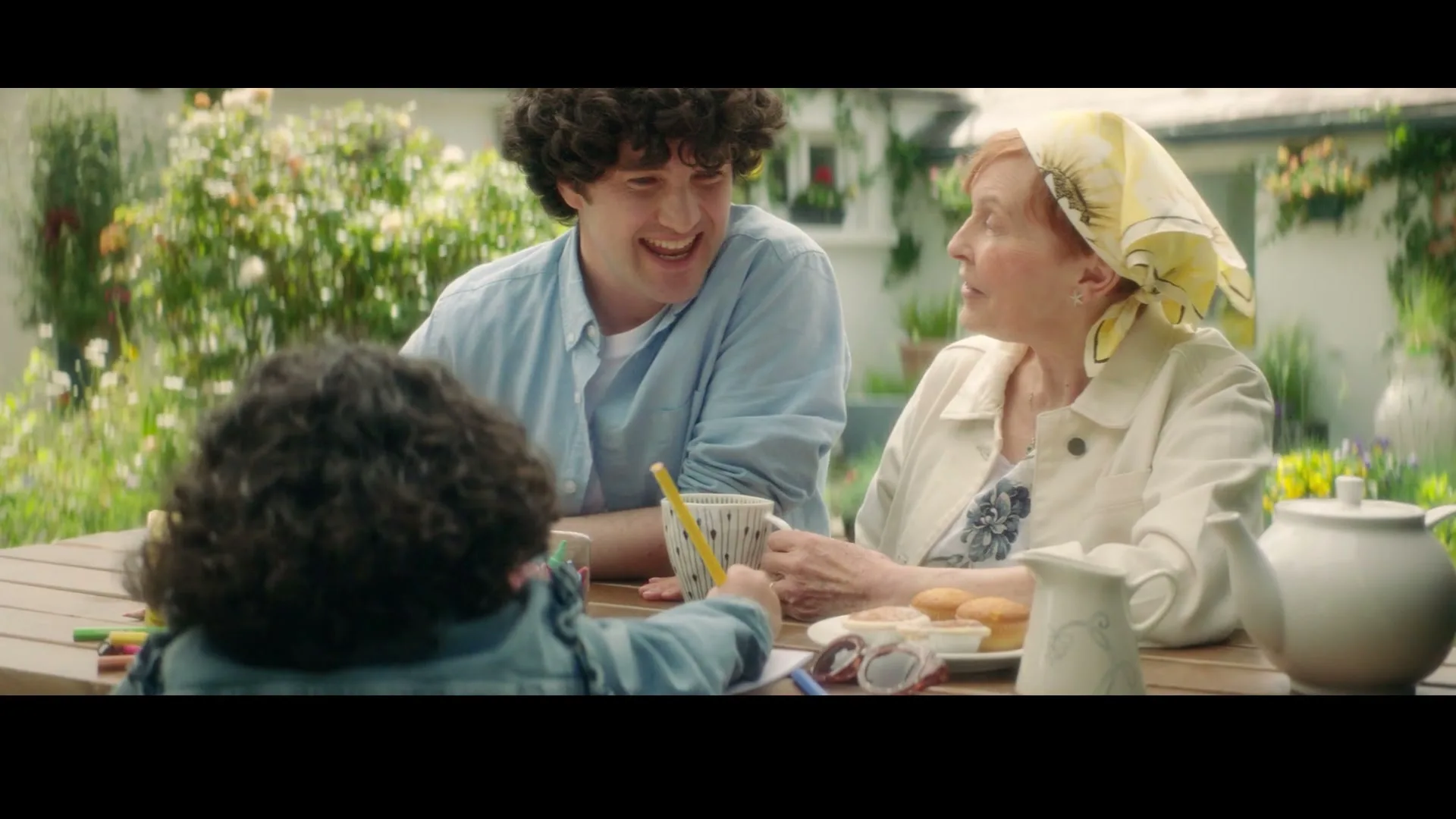 A young man and an older woman sit at a wooden table outdoors, smiling and looking at a child whose back is to the camera, in a sunny garden.