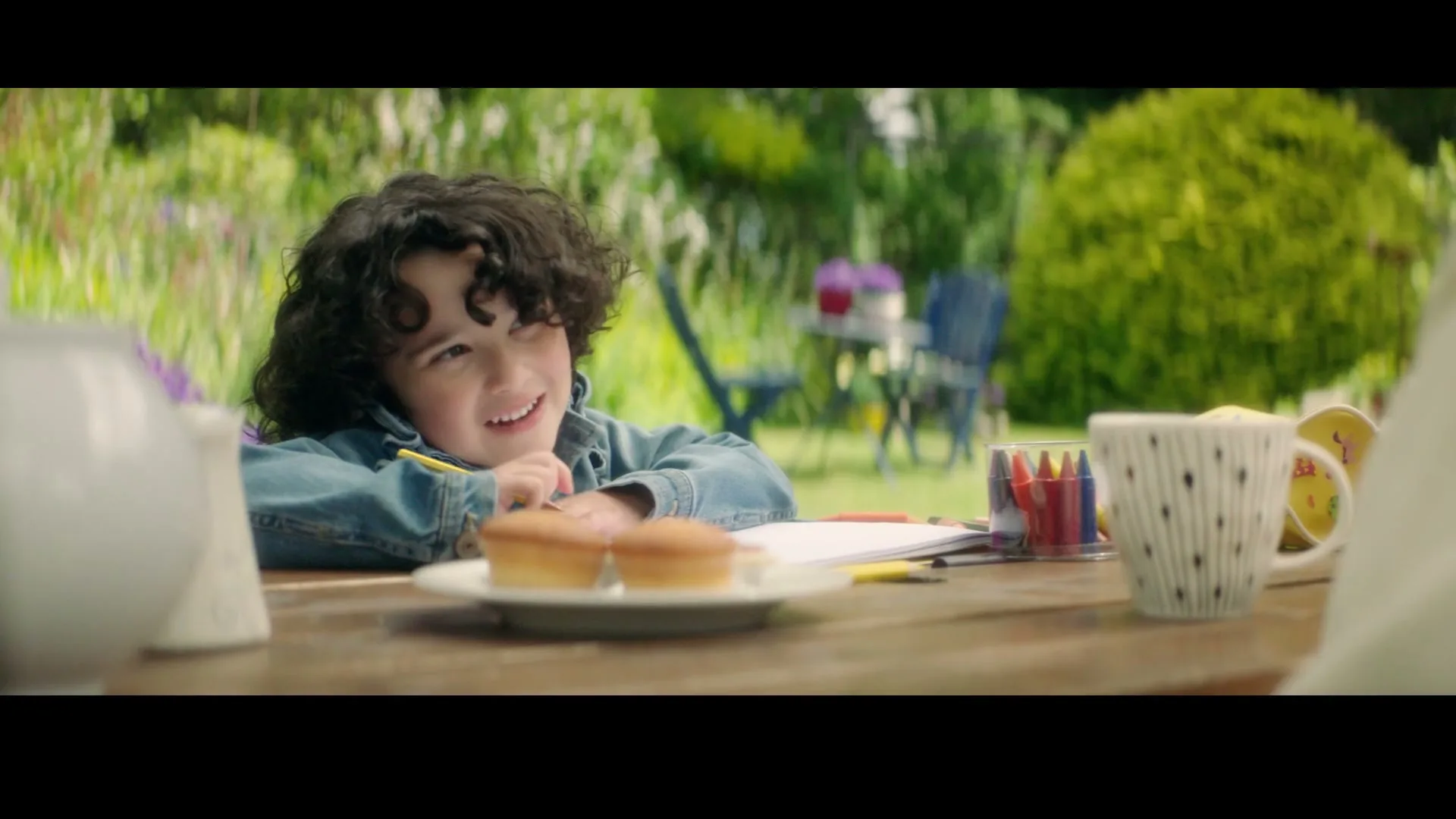 A young child with curly hair, wearing a denim jacket, sits at a wooden table in a garden, drawing and smiling.