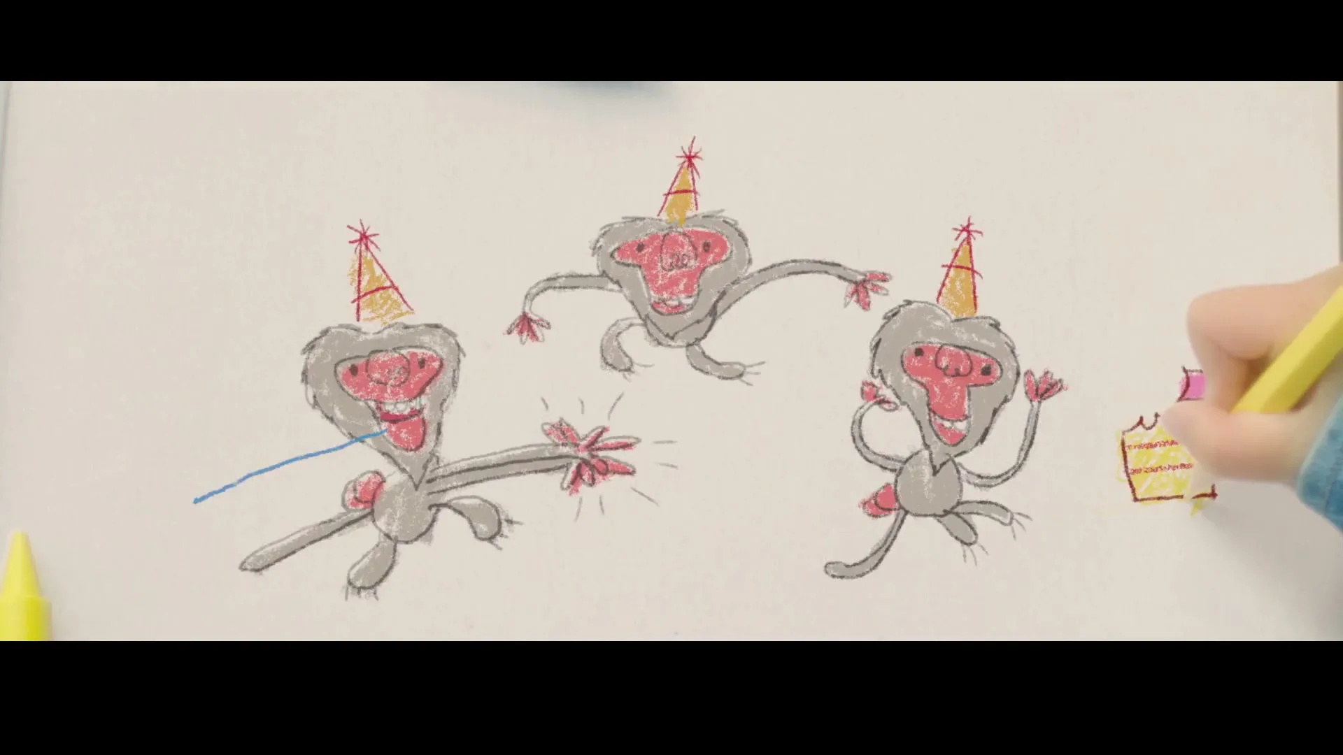 A child's hands are seen coloring a drawing of three monkeys wearing party hats and a slice of cake on a wooden table with crayons and sunglasses.