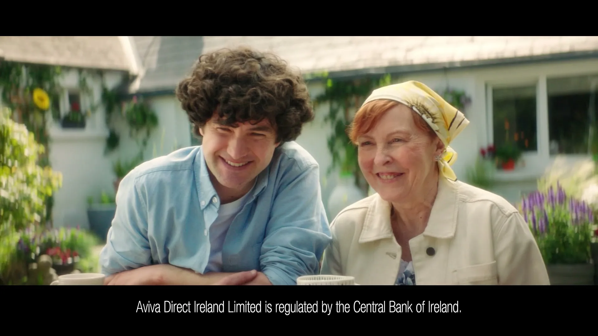 A young man with curly hair and an older woman with a headscarf sit at a table, smiling and looking at each other in a sunny garden.
