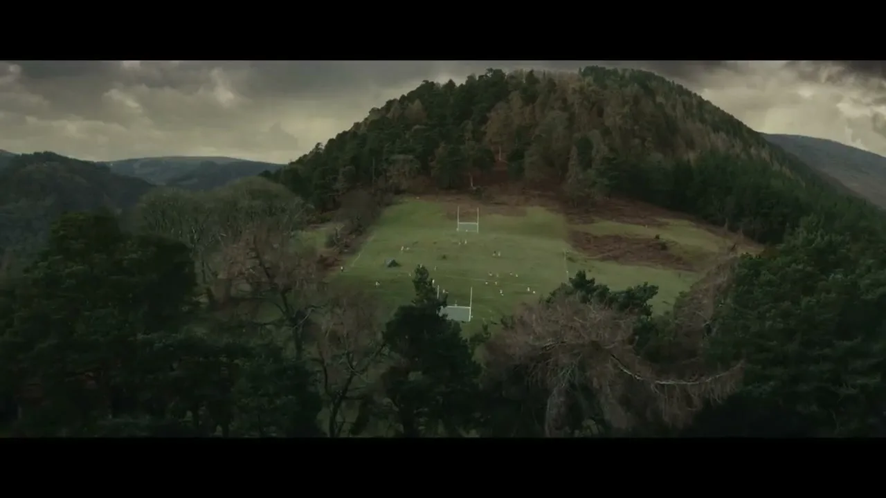 An aerial view of a small, green rugby field nestled in a valley surrounded by dark, forested hills under a cloudy sky.