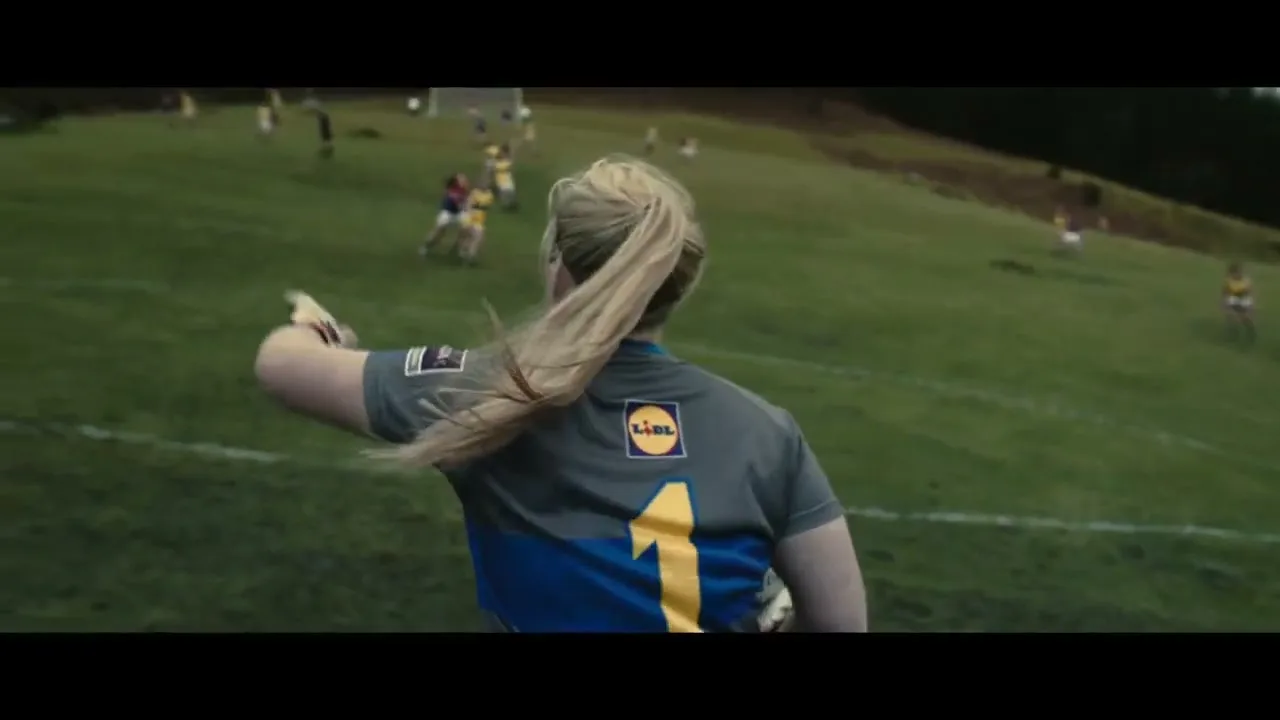 A female athlete with a blonde ponytail, wearing a grey and blue jersey with the number 1 and a Lidl logo, runs on a green field with other players in the background.