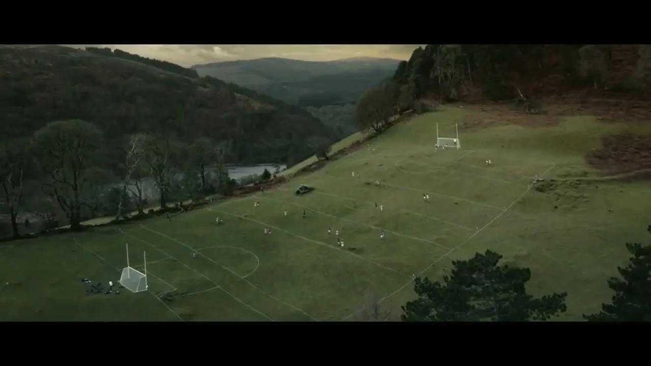 An aerial view shows a women's Gaelic football match in progress on a green field nestled in a valley with a lake and hills.