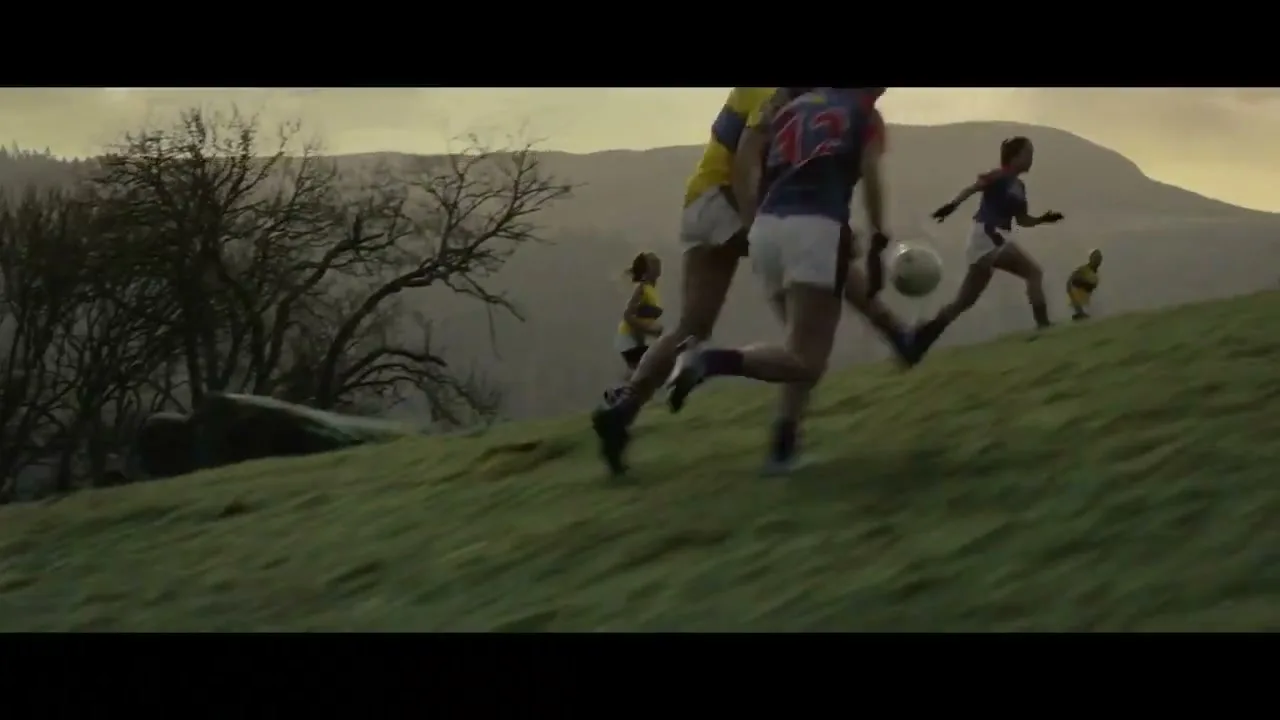 A female Gaelic football player in a yellow jersey catches the ball on a grassy field with other players and hills in the background.
